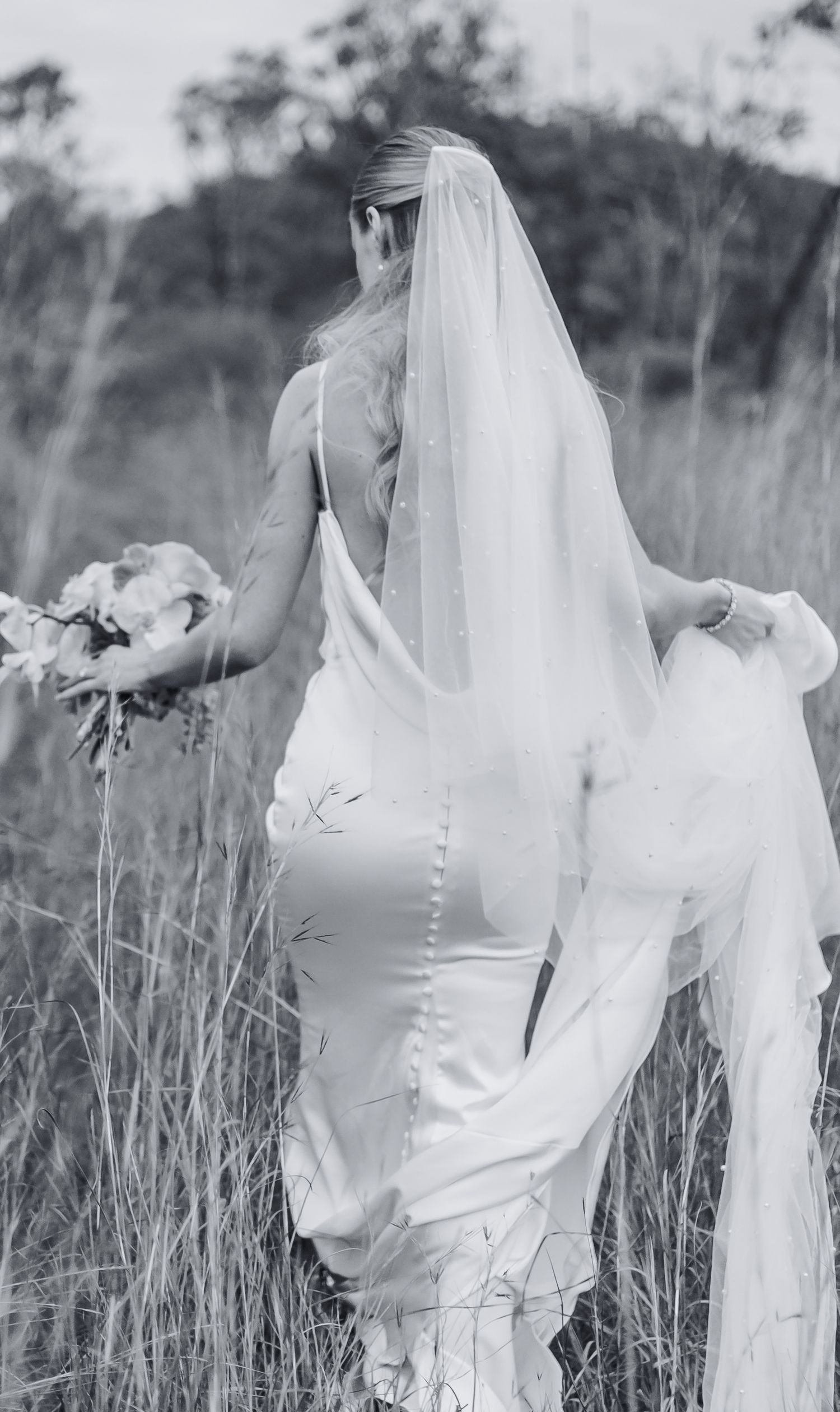 Bride in fitted white dress with veil walking through tall grass in outdoor setting.