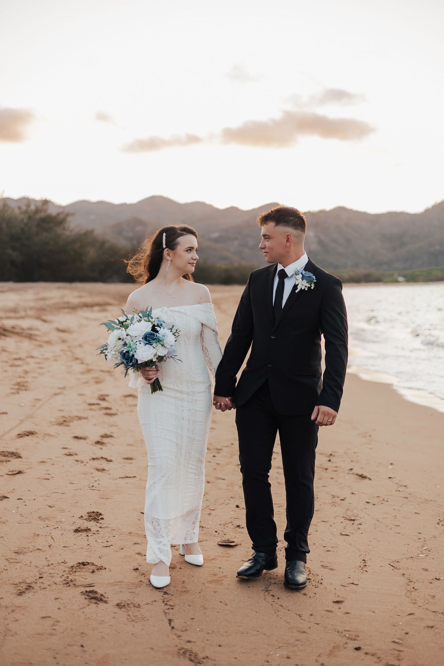 Newlyweds stroll along beach at sunset with bride holding blue and white bouquet.