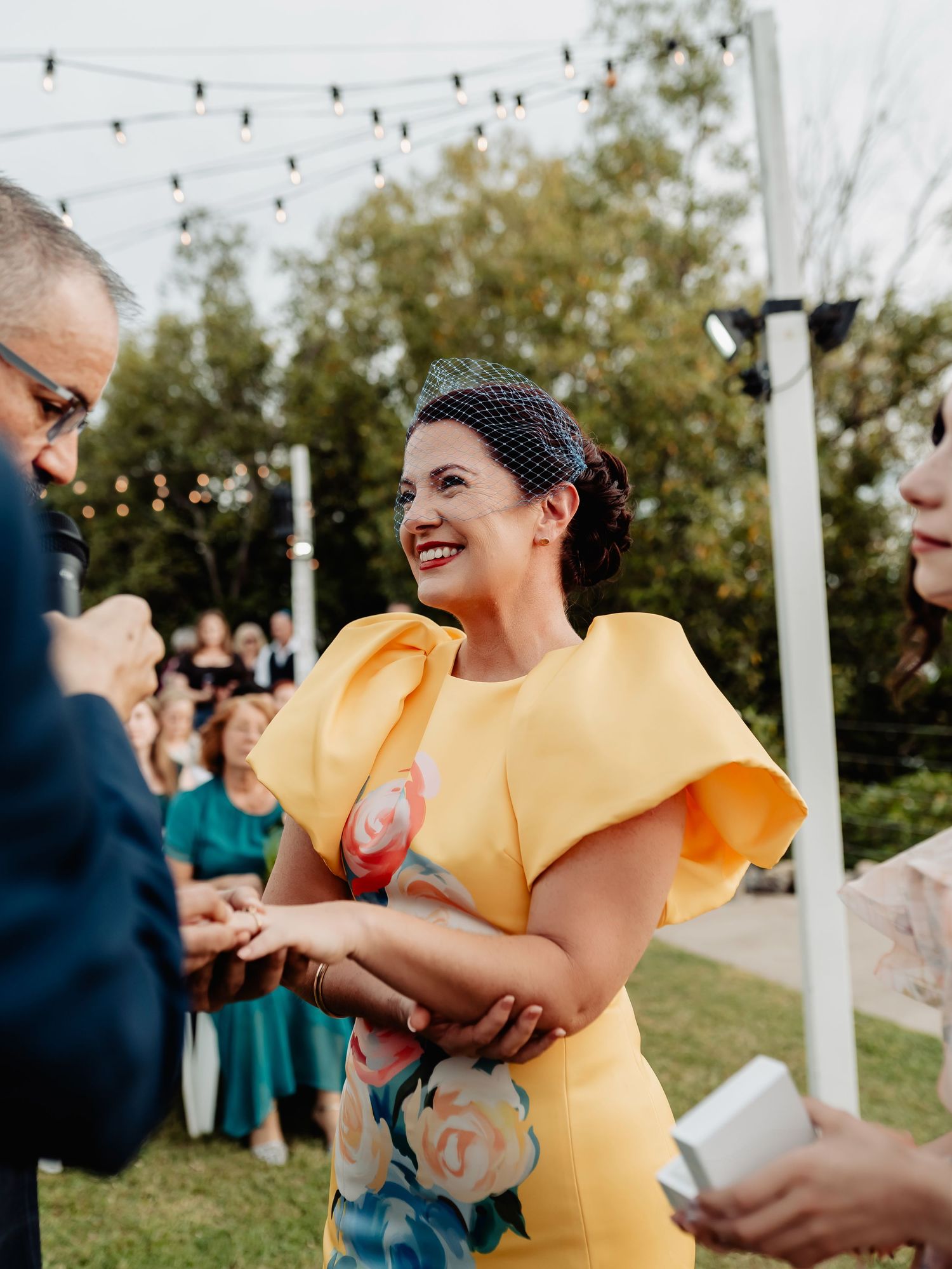 Woman in yellow floral dress with ruffled sleeves smiling at outdoor wedding ceremony with string lights.