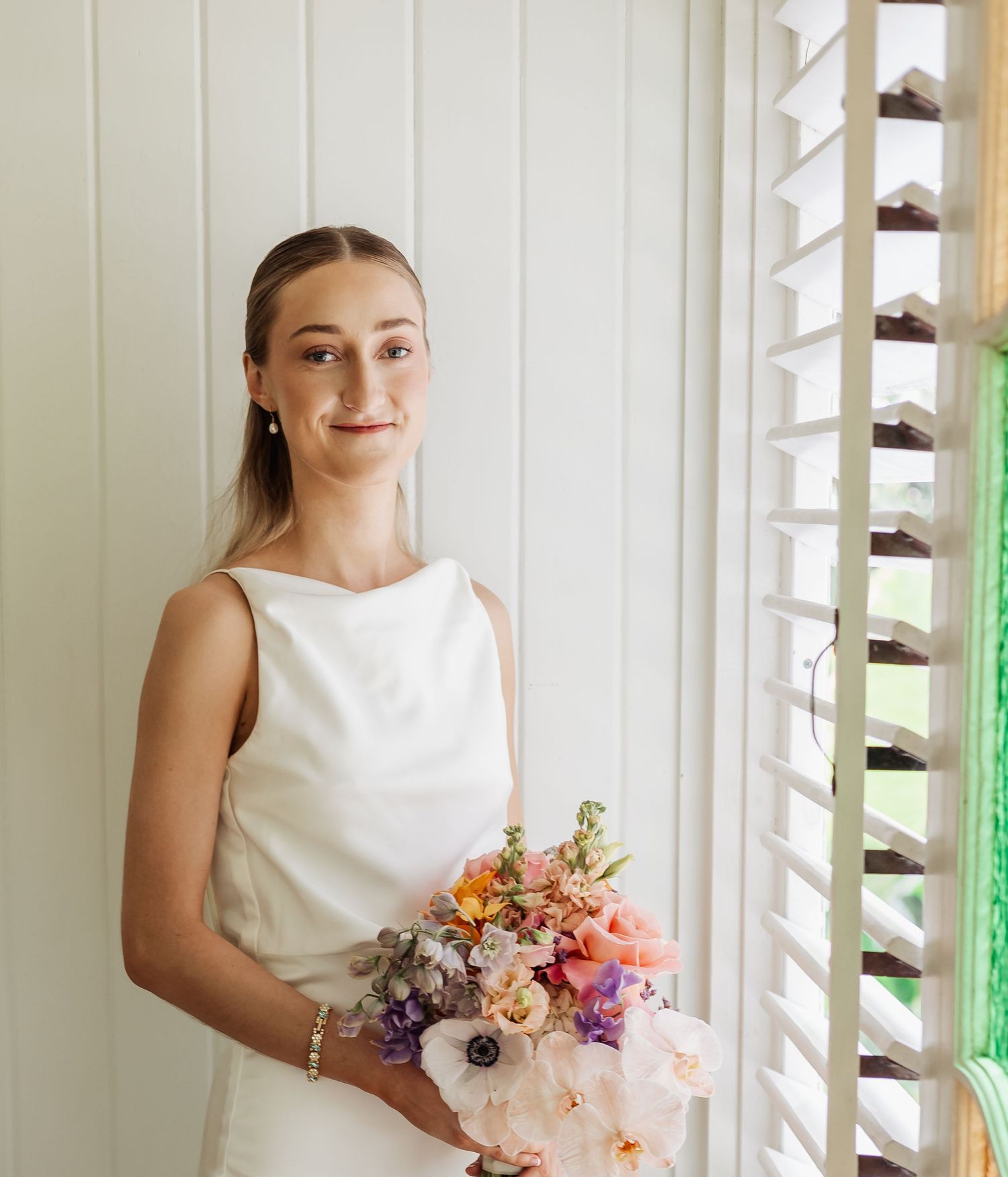 Bride holding colorful bouquet poses in front of white shuttered wall.