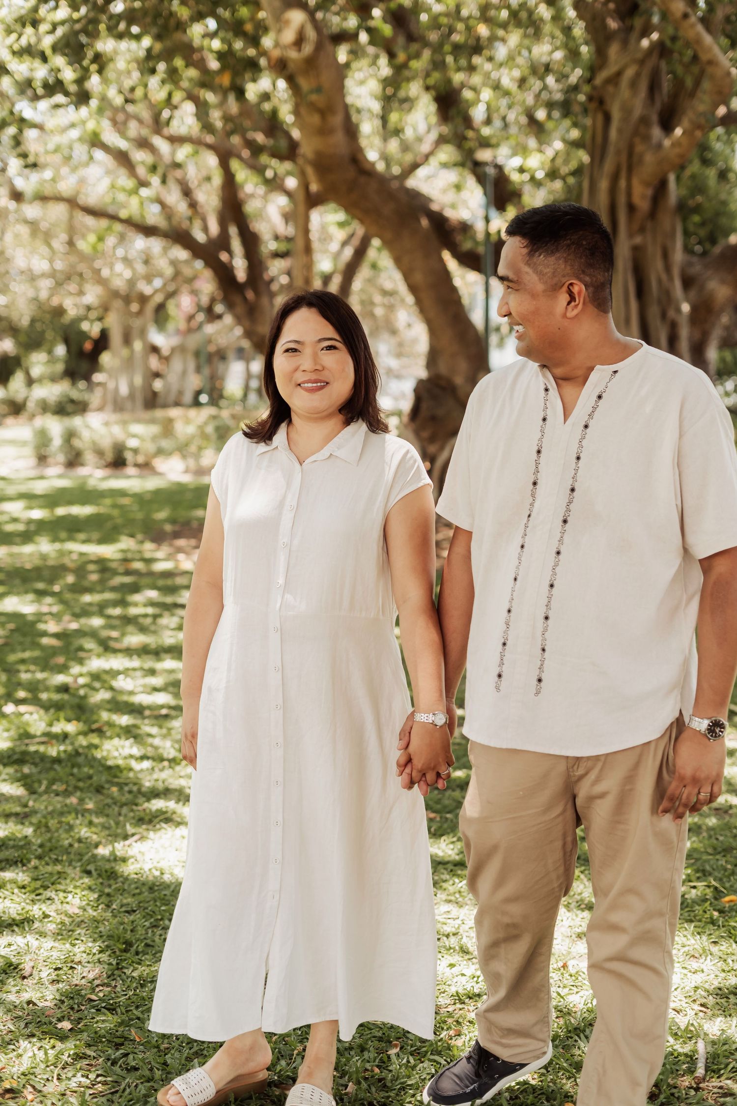 A family dressed in coordinating white and beige outfits poses together under large trees in a park.
