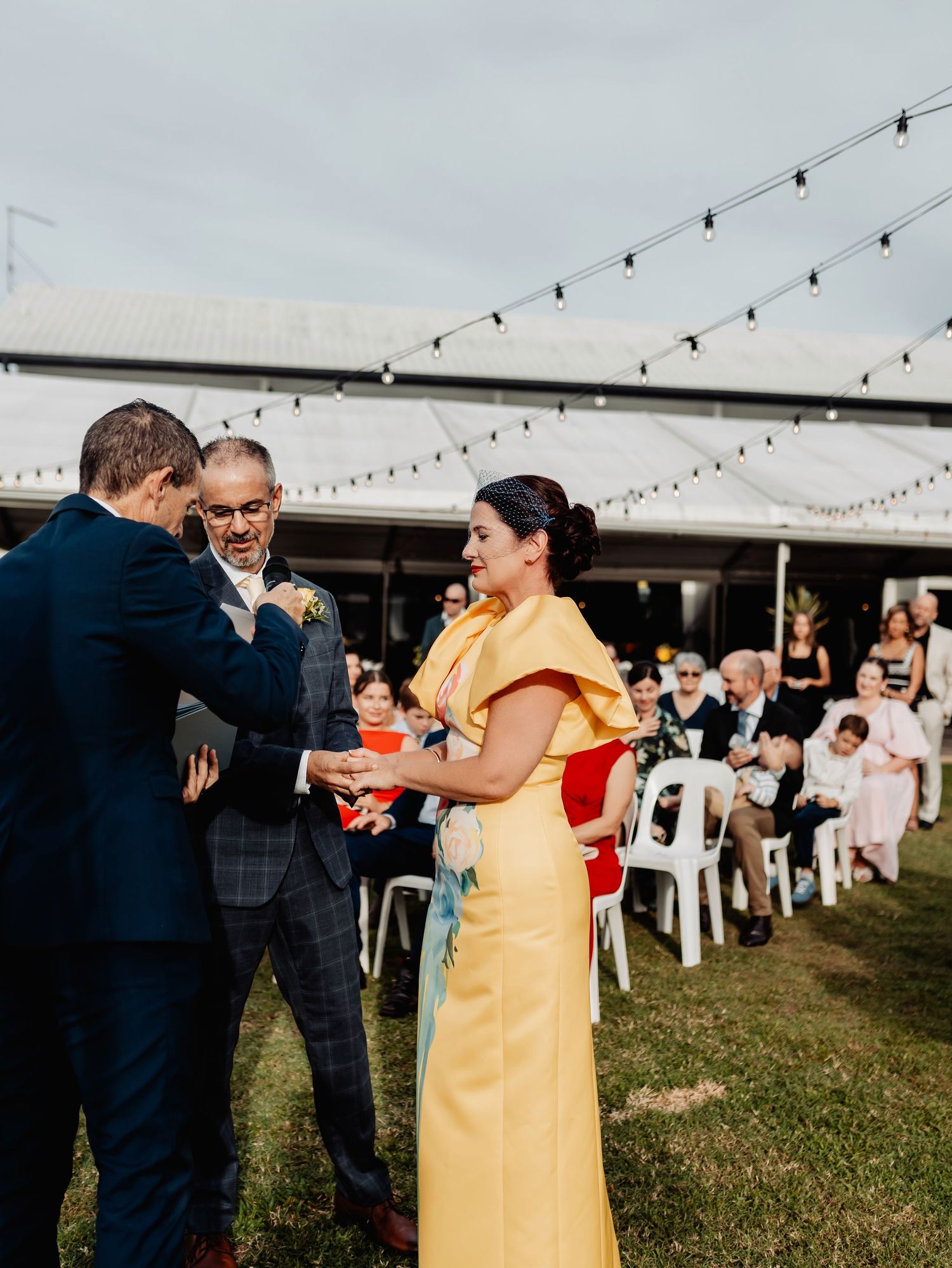 Woman in yellow gown greeting guests at an outdoor wedding ceremony with string lights overhead.
