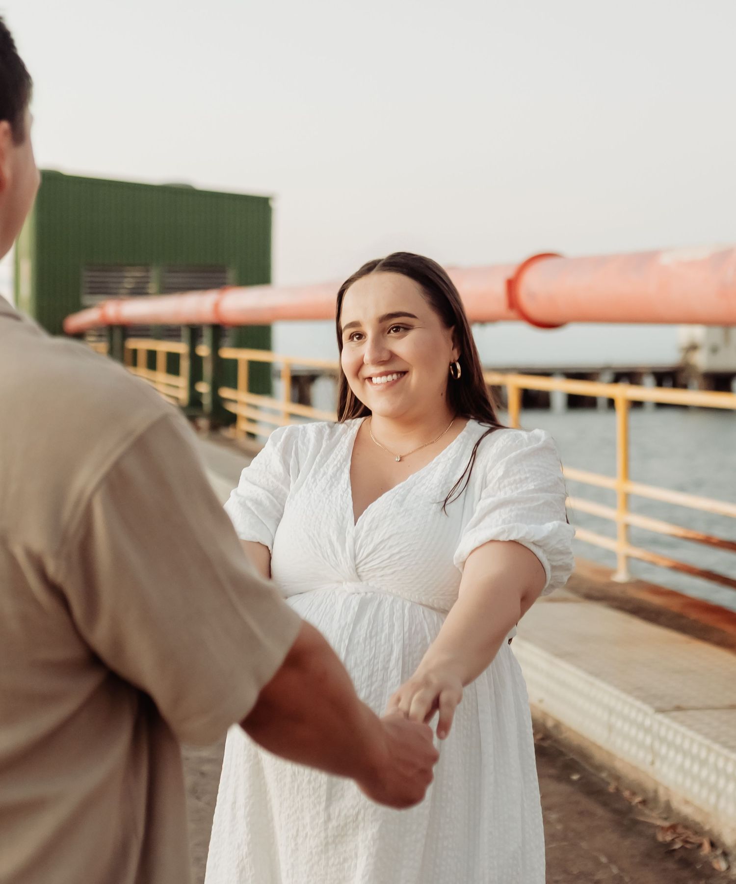 A smiling woman in a white dress holds hands with a man on a waterfront pier at golden hour.