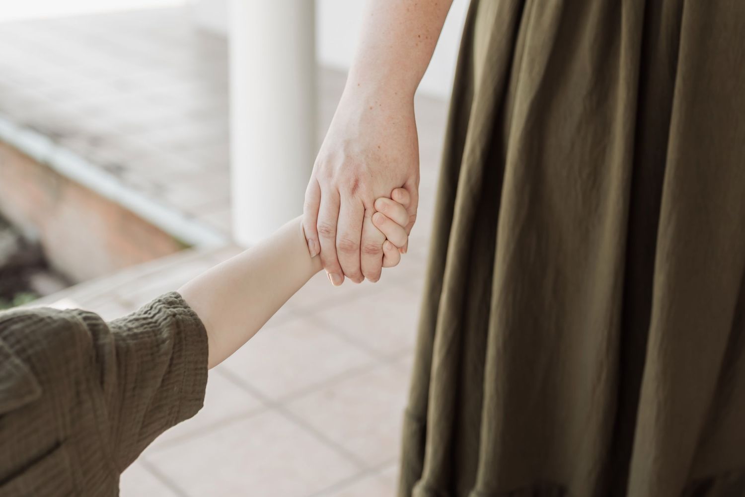 Adult and child holding hands tenderly, wearing olive green clothing, in a warm indoor setting.