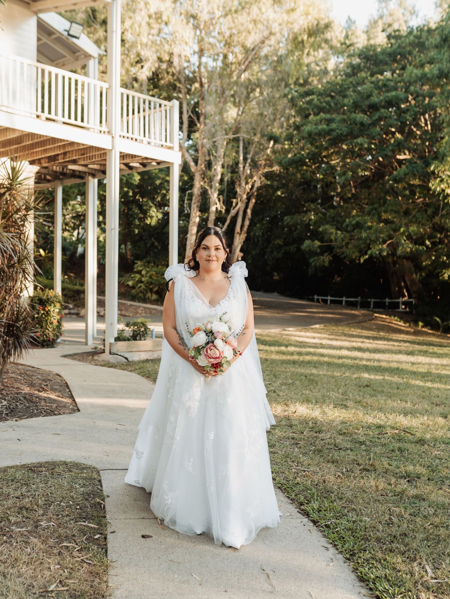 Bride in white floral gown holds bouquet on garden path beside white colonial house.