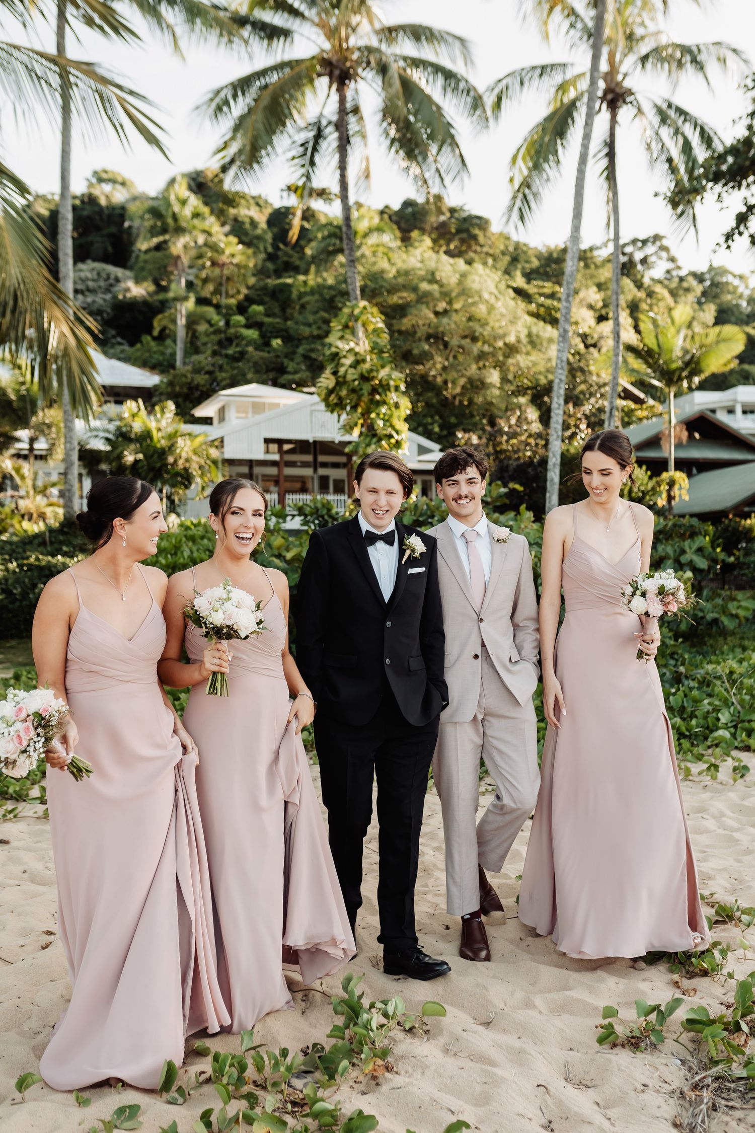 Wedding party walks on sandy beach, groom in tuxedo flanked by bridesmaids in blush gowns and groomsman in gray suit.