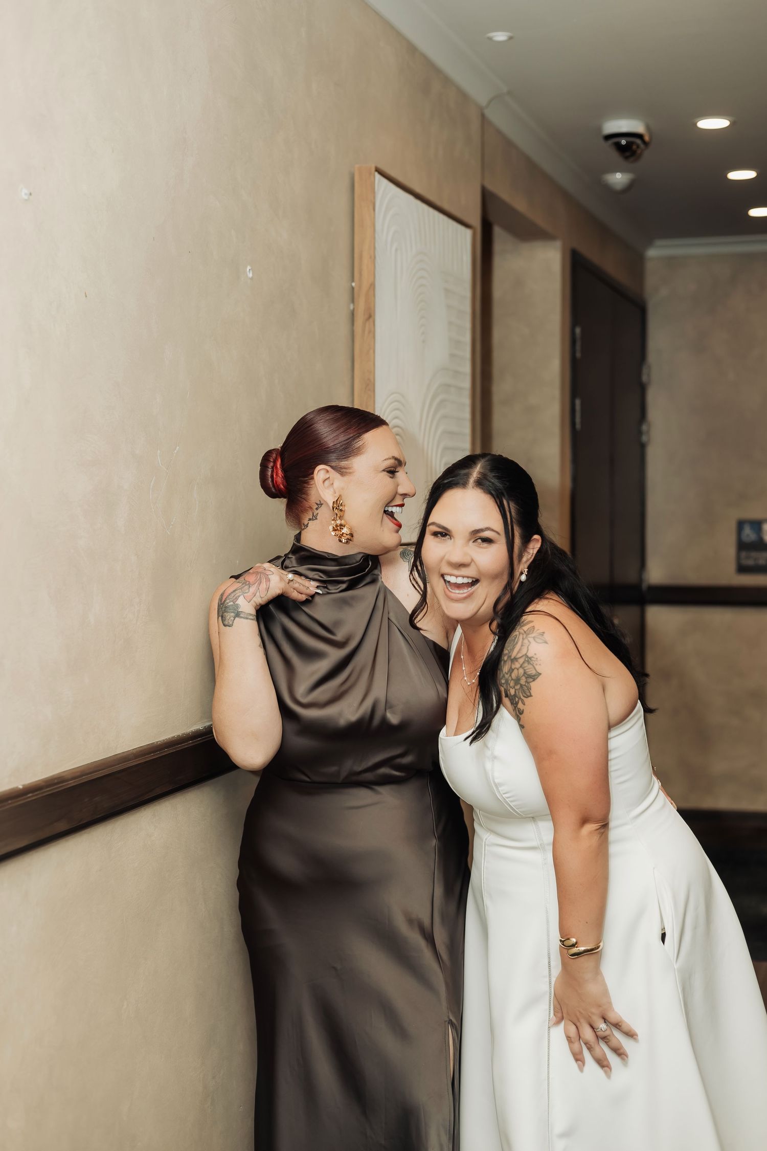 Two women laughing together at a wedding, one in a white bridal gown and one in a grey satin dress.