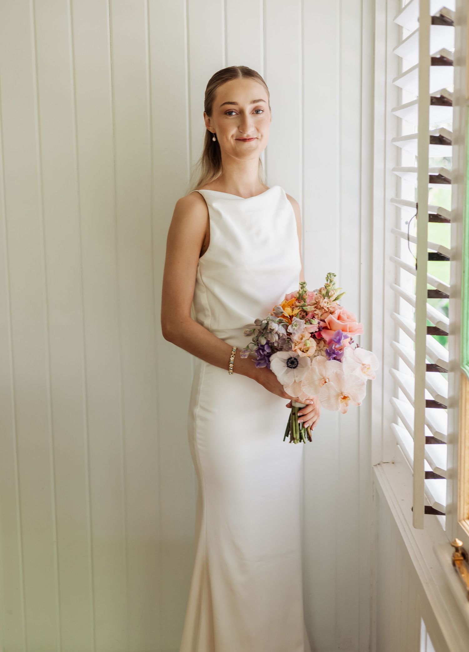 Elegant bride in sleek white gown poses by window with natural light streaming in.