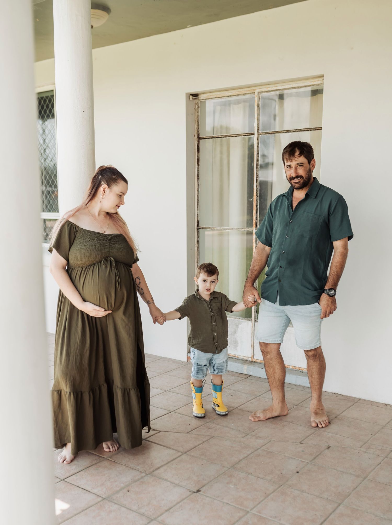 Pregnant woman in olive dress with toddler and man on tiled porch, family maternity photo outdoors.