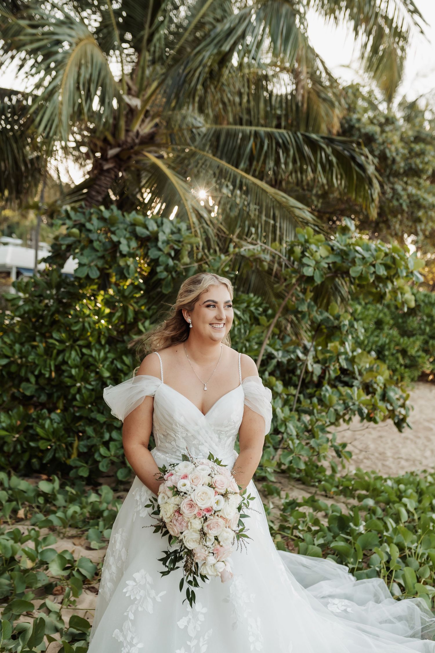 Bride in off-shoulder white gown holds pink bouquet, smiling outdoors among tropical greenery and palm trees.
