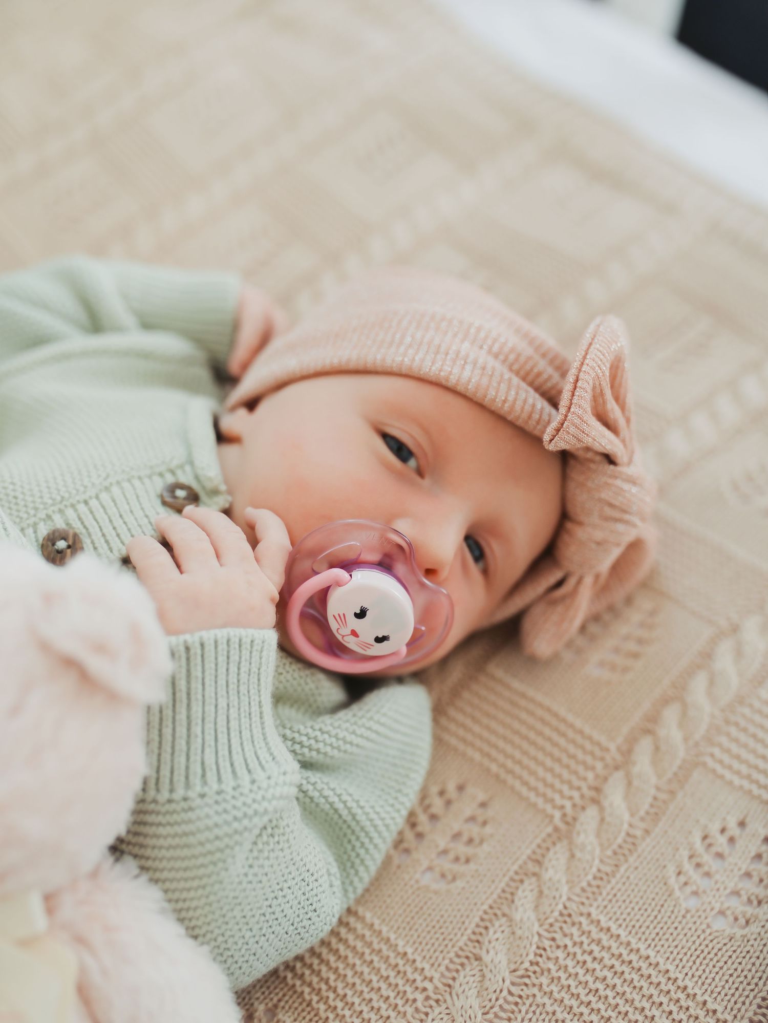 Newborn baby wearing a mint green knit cardigan and pink bow hat, lying on a beige blanket with a pacifier.