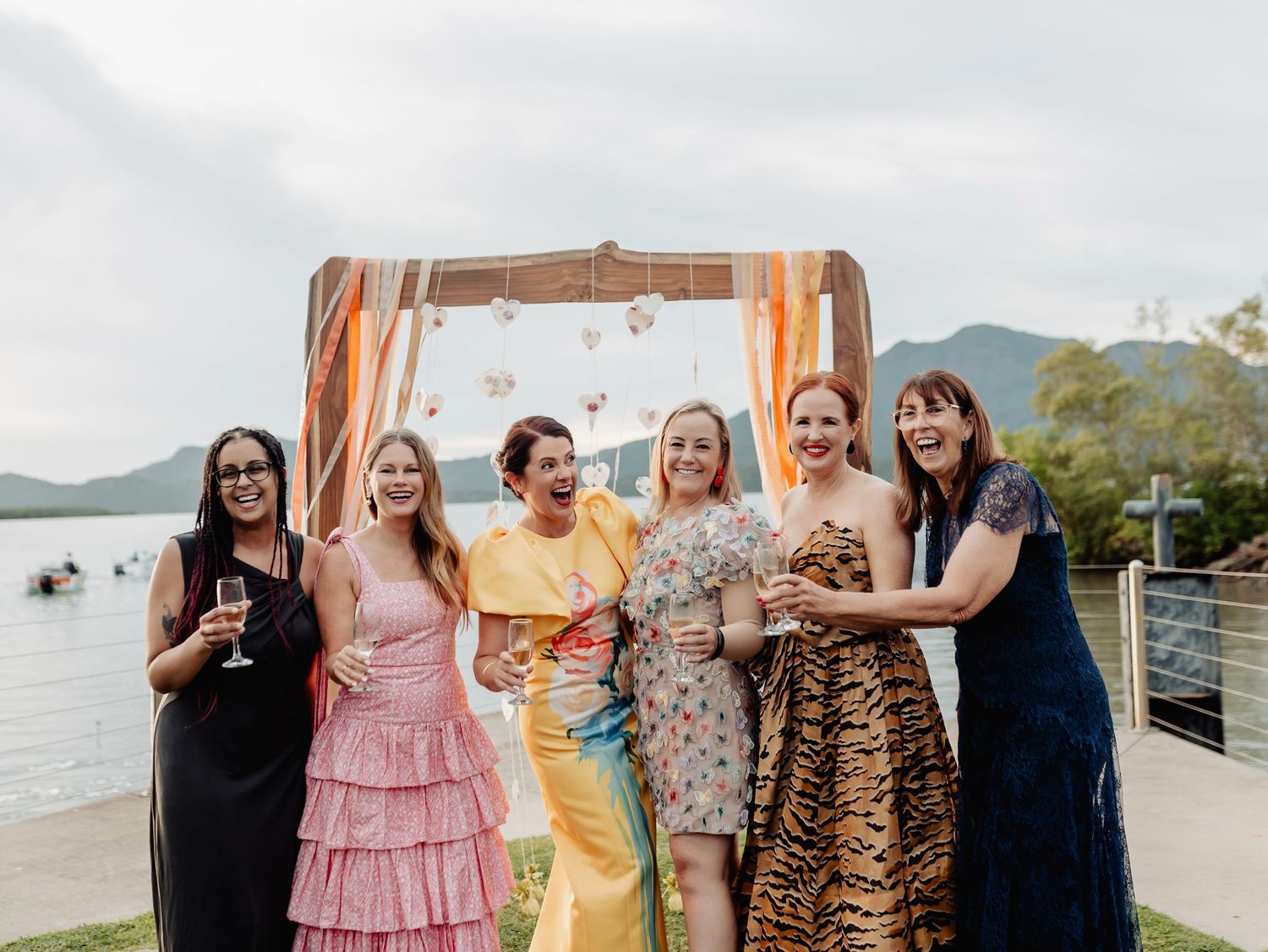 Six women in colorful dresses laughing and celebrating outdoors near a waterfront wedding arch.