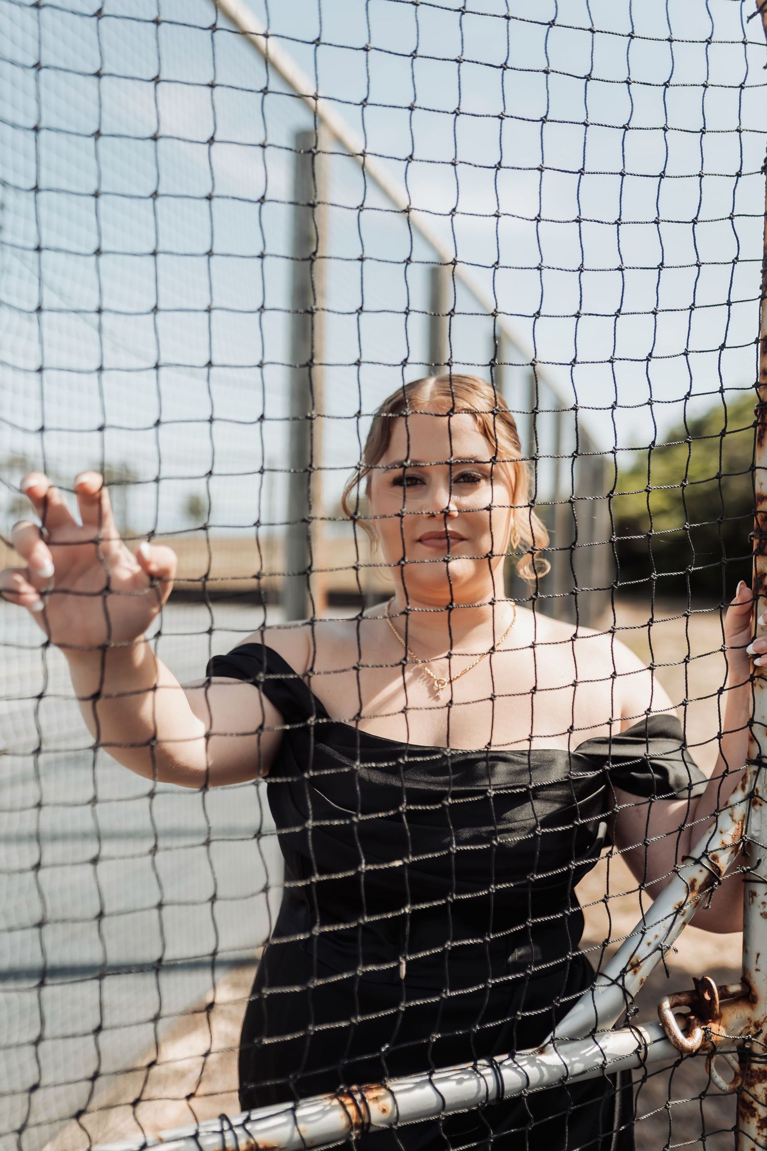 A person in black poses playfully behind a mesh fence against a bright sky background.