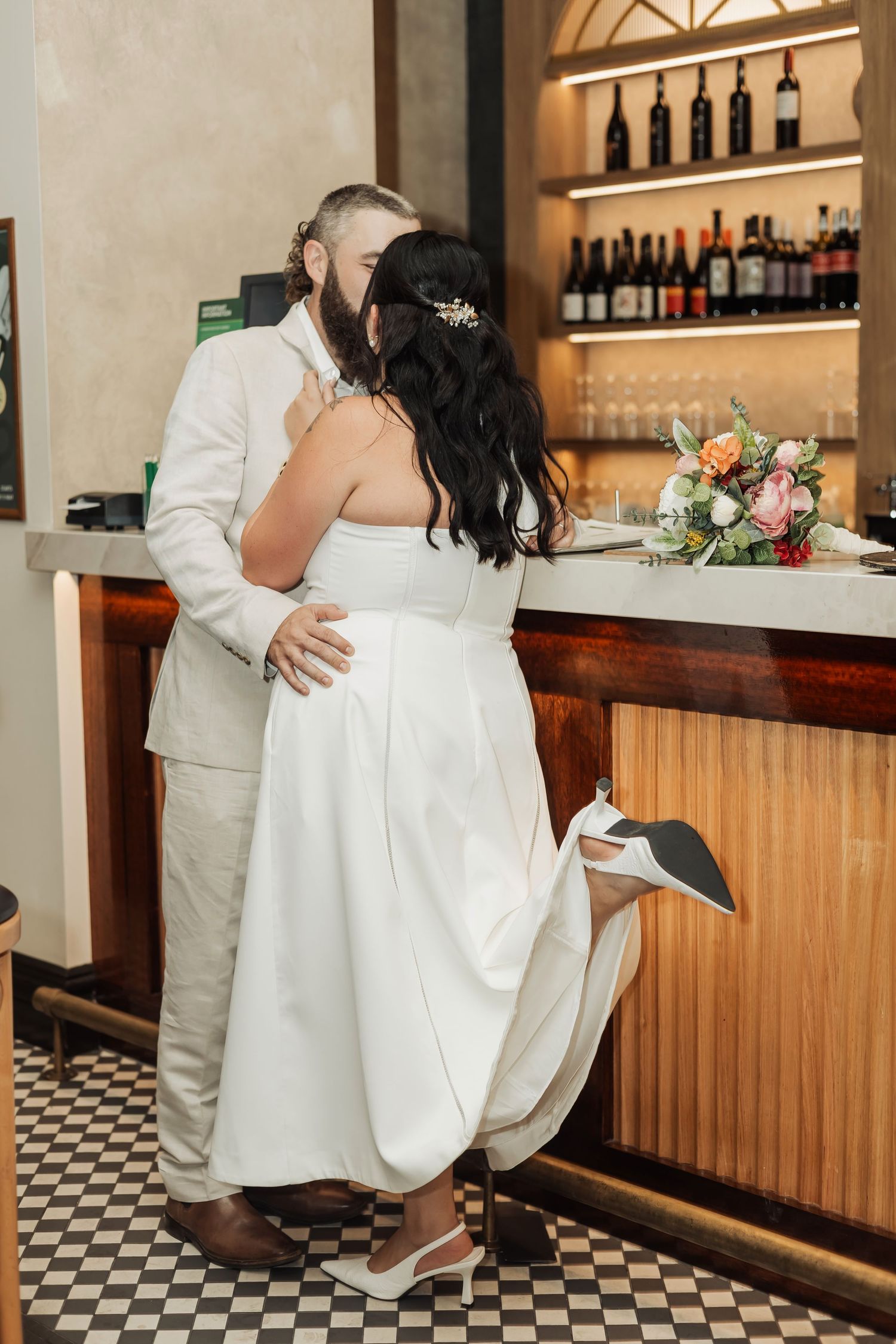 Newlyweds share a romantic kiss at a bar, bride in white gown with bouquet nearby, groom in light suit.