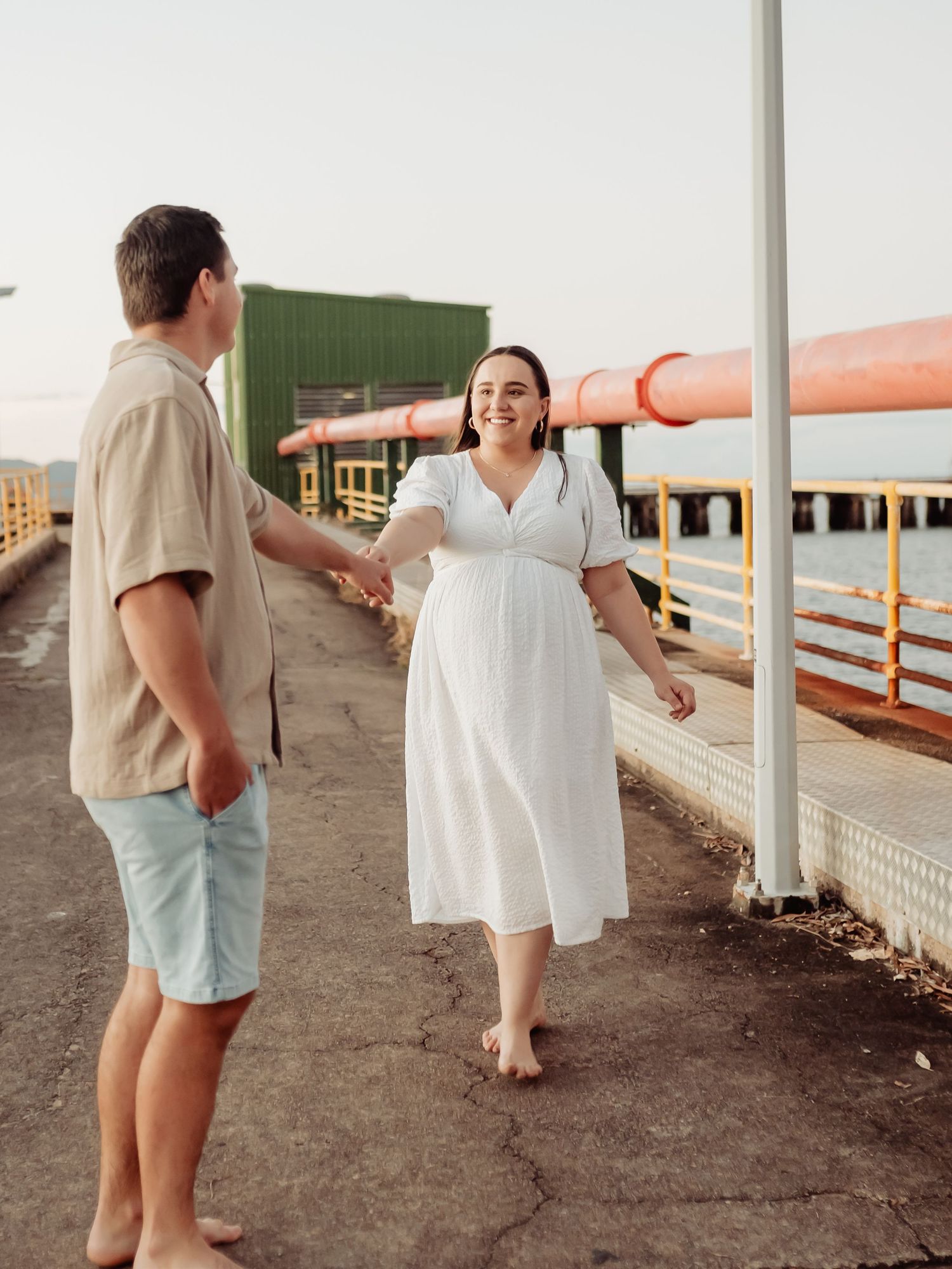 Couple holding hands on a waterfront pier, woman in white dress and man in casual shorts, golden hour light.