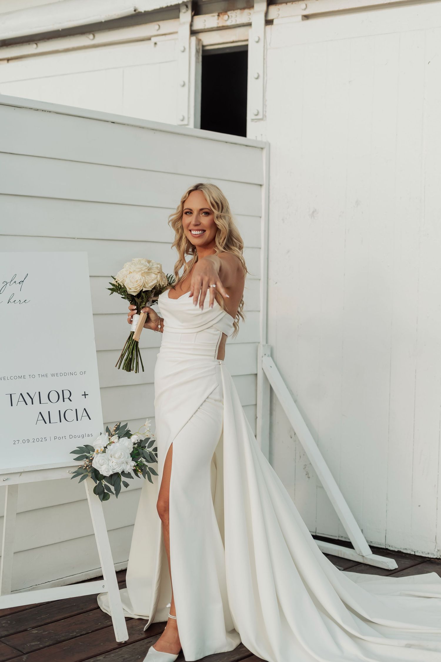 Bride in white dress poses playfully in front of white wall with wedding signage.