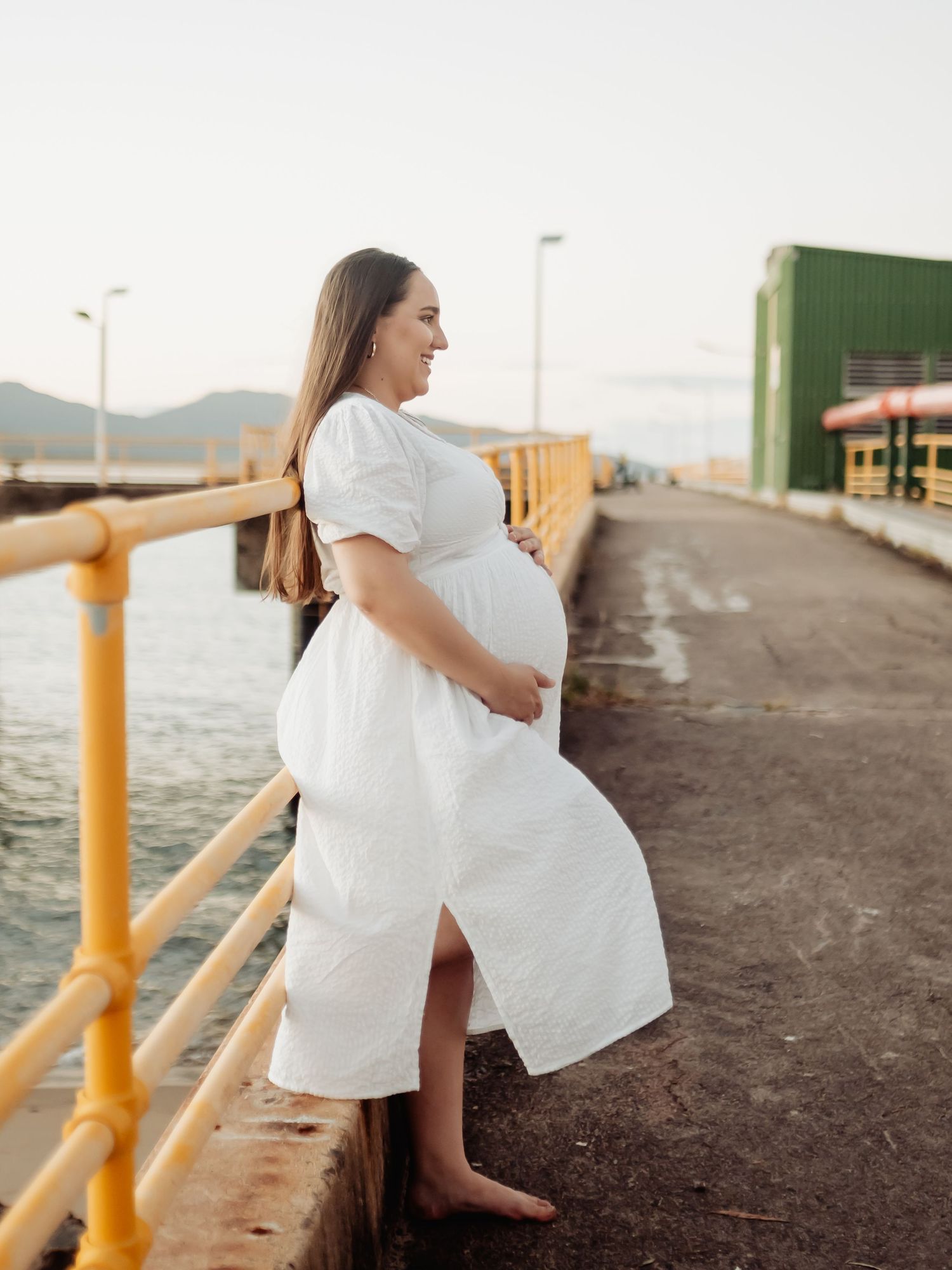 Pregnant woman in white dress holding belly on waterfront pier at sunset.