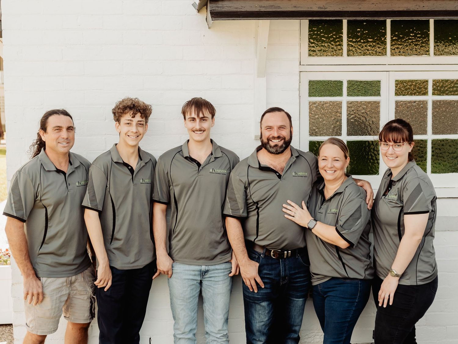 Six team members in matching grey polo shirts smiling together outside a white building.