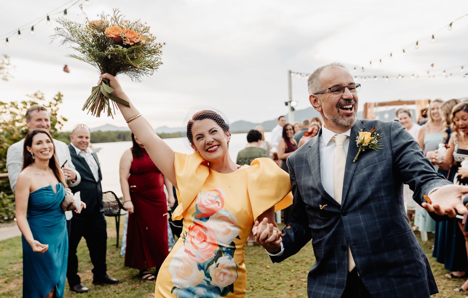 Joyful couple celebrating wedding exit with colorful bouquet raised high, guests cheering outdoors near waterfront.