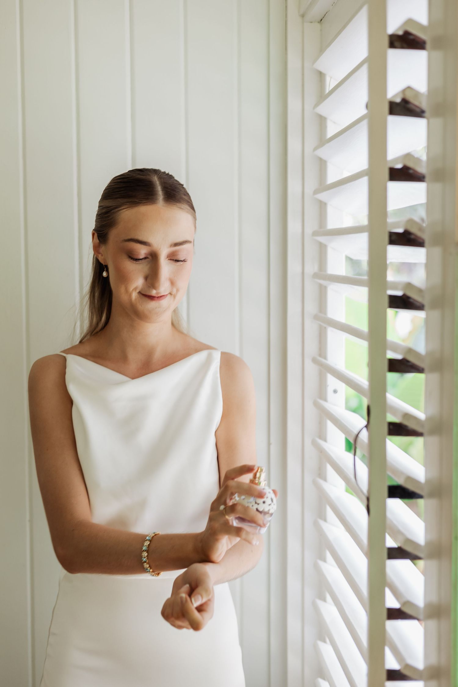 Elegant bride in white sleeveless dress preparing in front of shuttered windows.