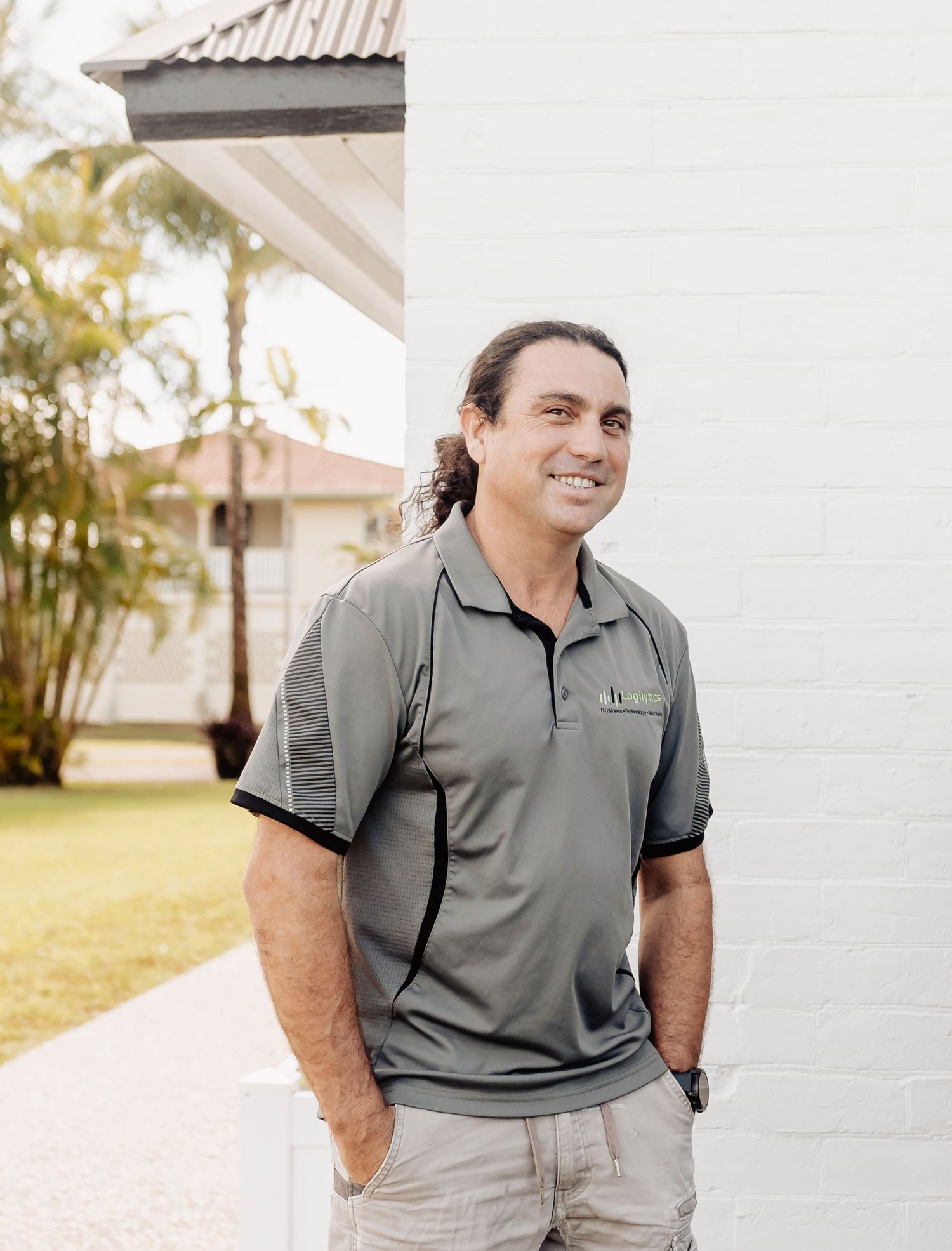 Smiling man with long hair in gray polo shirt leaning against white brick wall outdoors.
