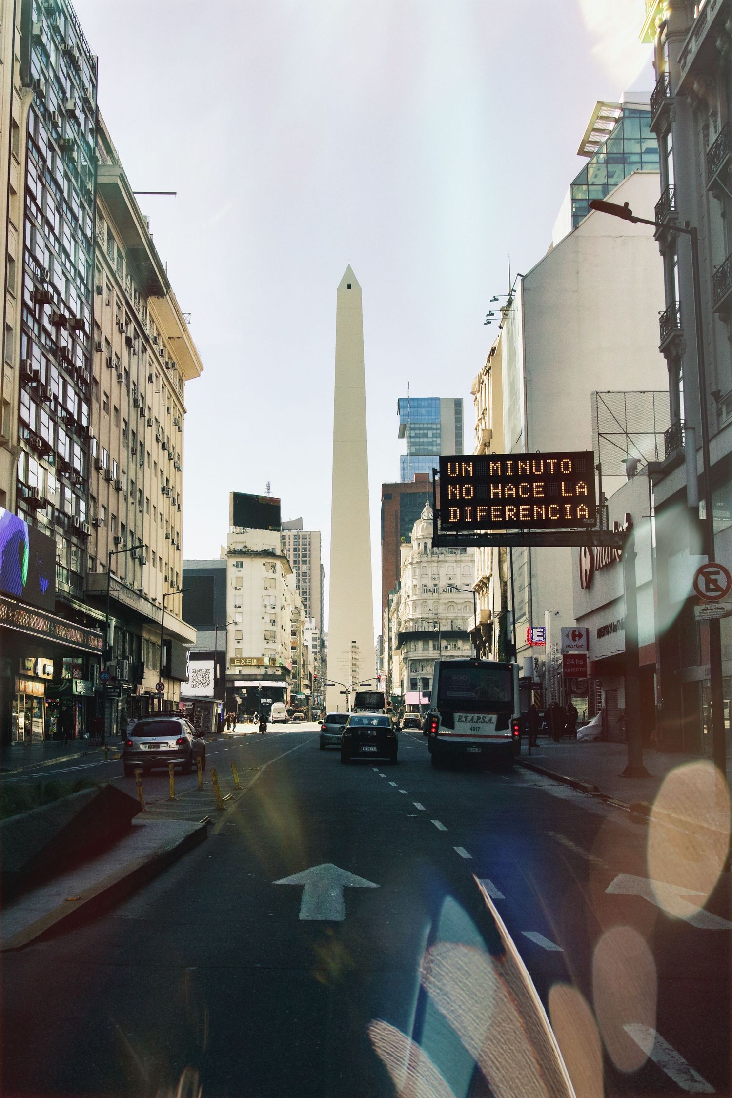 Urban street view showing the iconic Obelisk monument in downtown Buenos Aires against a bright sky.