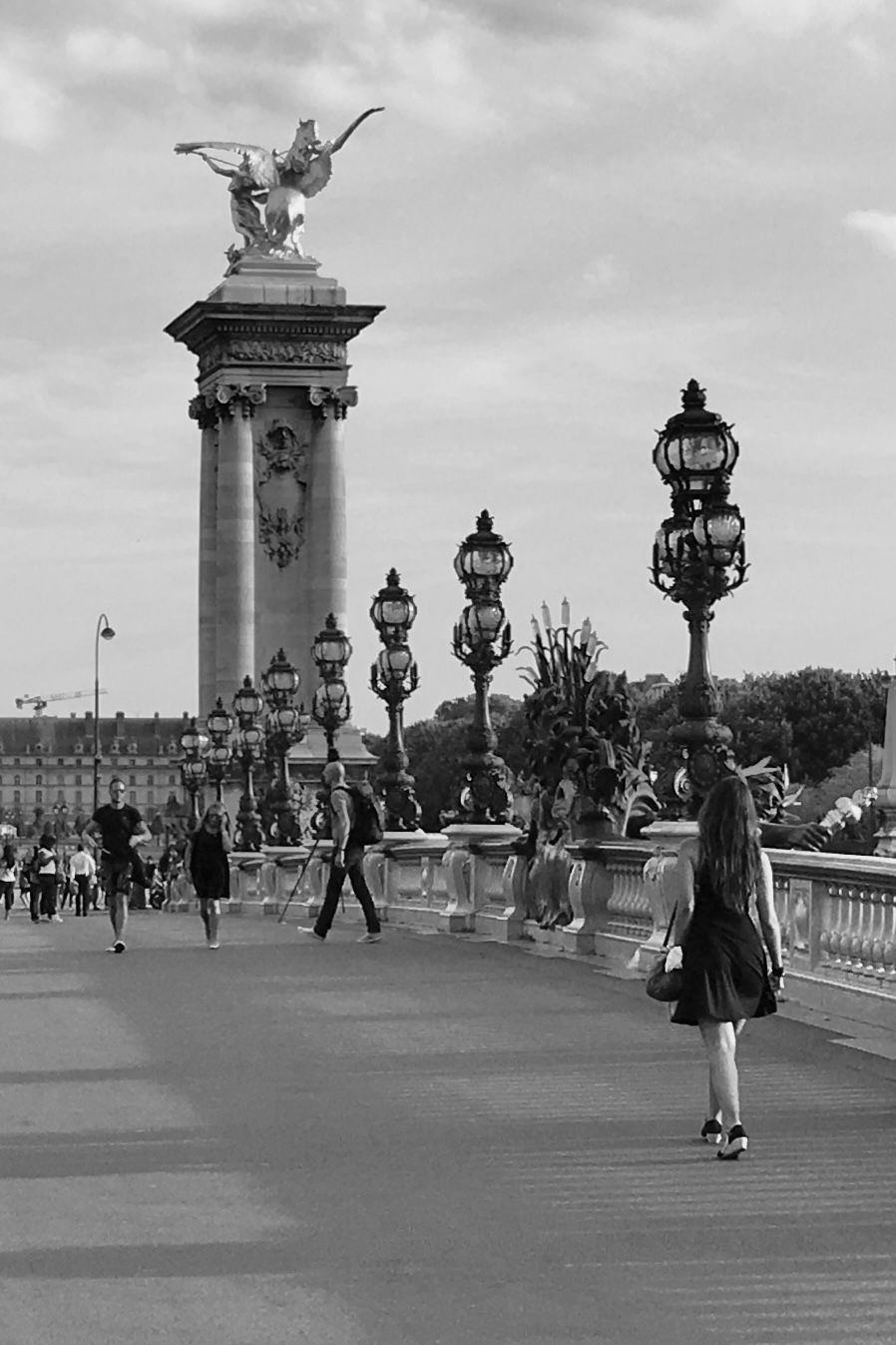 Black and white photograph of people strolling on the historic Pont Alexandre III bridge in Paris with ornate lampposts.