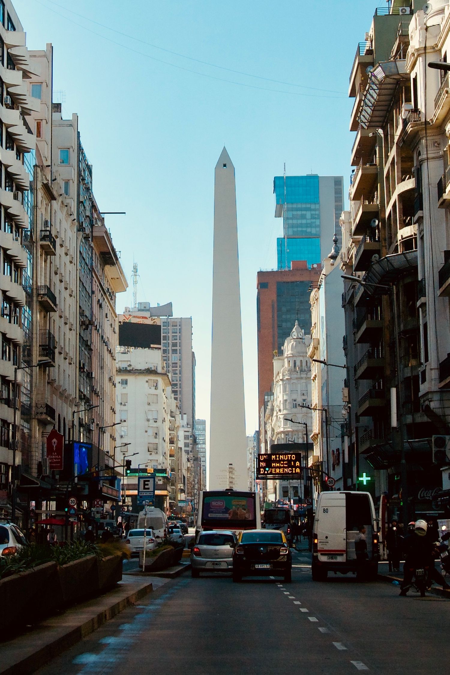 View down a Buenos Aires street with the iconic Obelisk monument visible in the distance between city buildings.