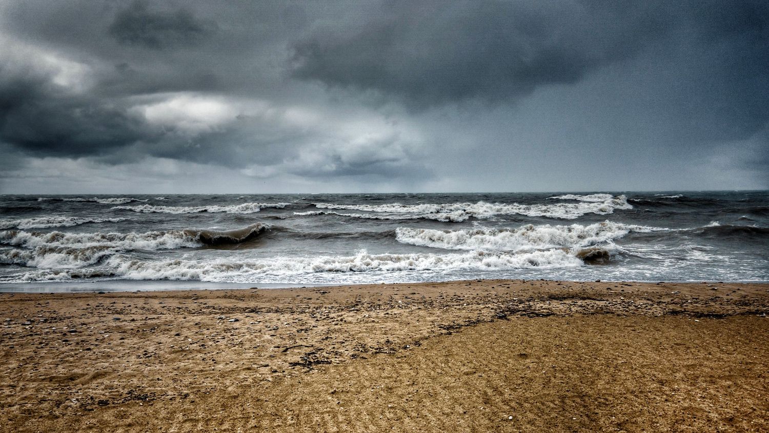 Dramatic stormy seascape with dark clouds looming over choppy waves and sandy beach.