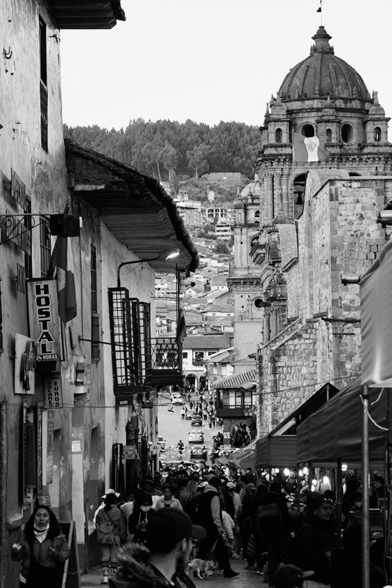 Busy street scene in Cusco, Peru with colonial architecture and domed church tower against hillside buildings in black and white.