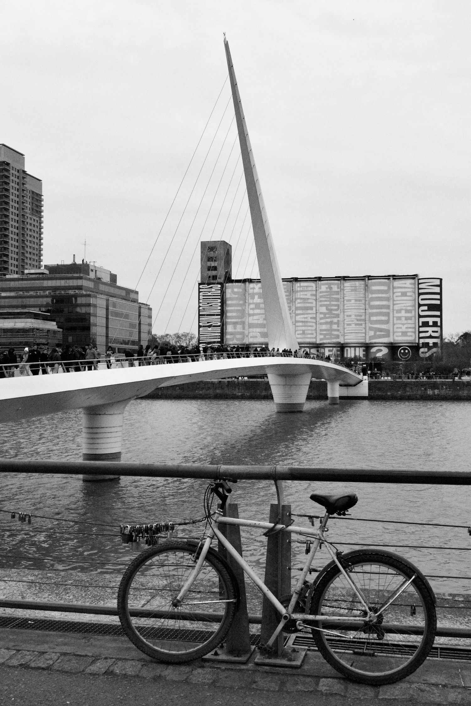 Black and white photograph of bicycles parked along a modern bridge over calm waters.