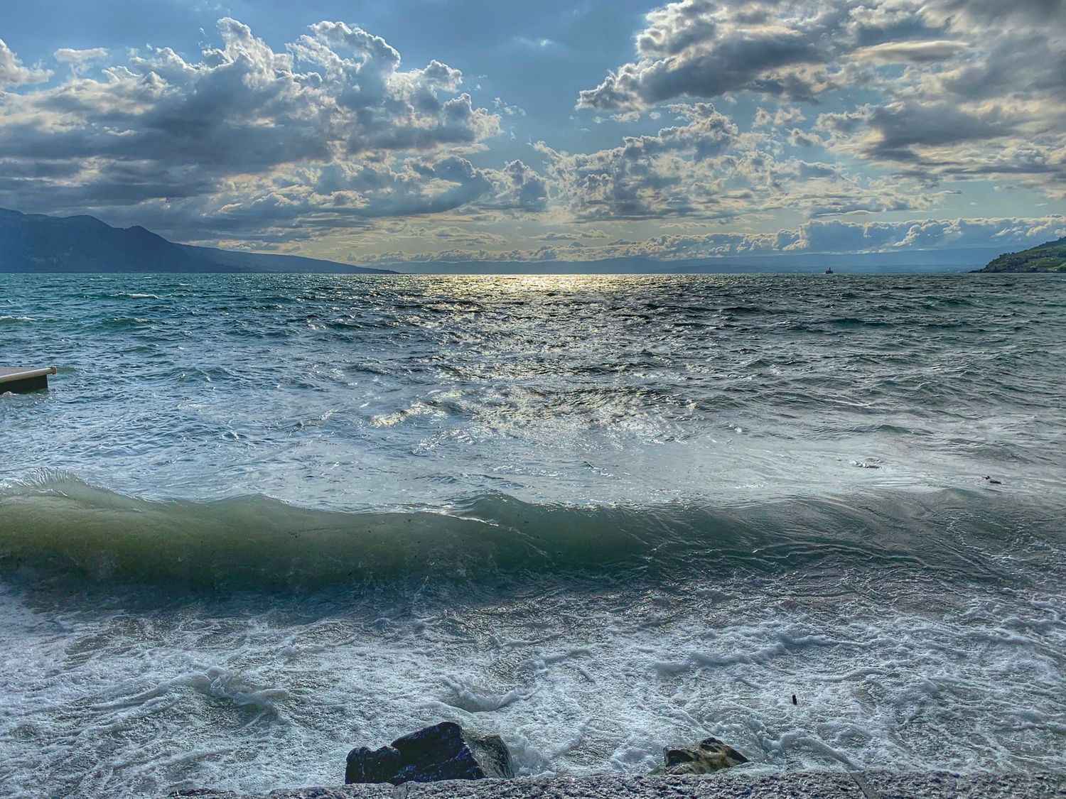 Dramatic cloudy sky over choppy ocean waves crashing against rocks with mountains in the distance.