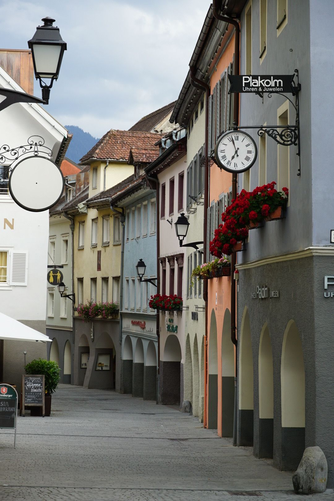 Historic European street with arched walkways, traditional lampposts, and red flower boxes decorating the building facades.