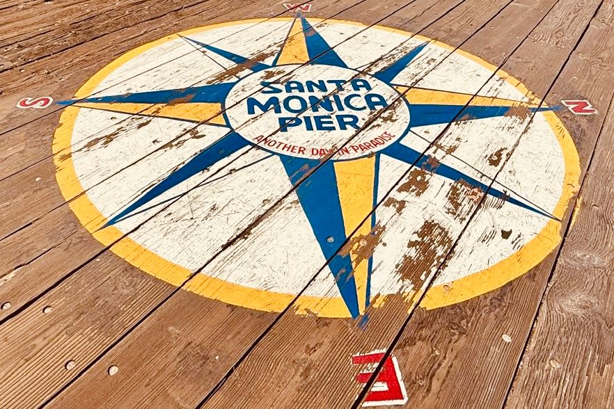 A decorative compass rose design inlaid on wooden deck planks features blue and yellow colors with directional markers.