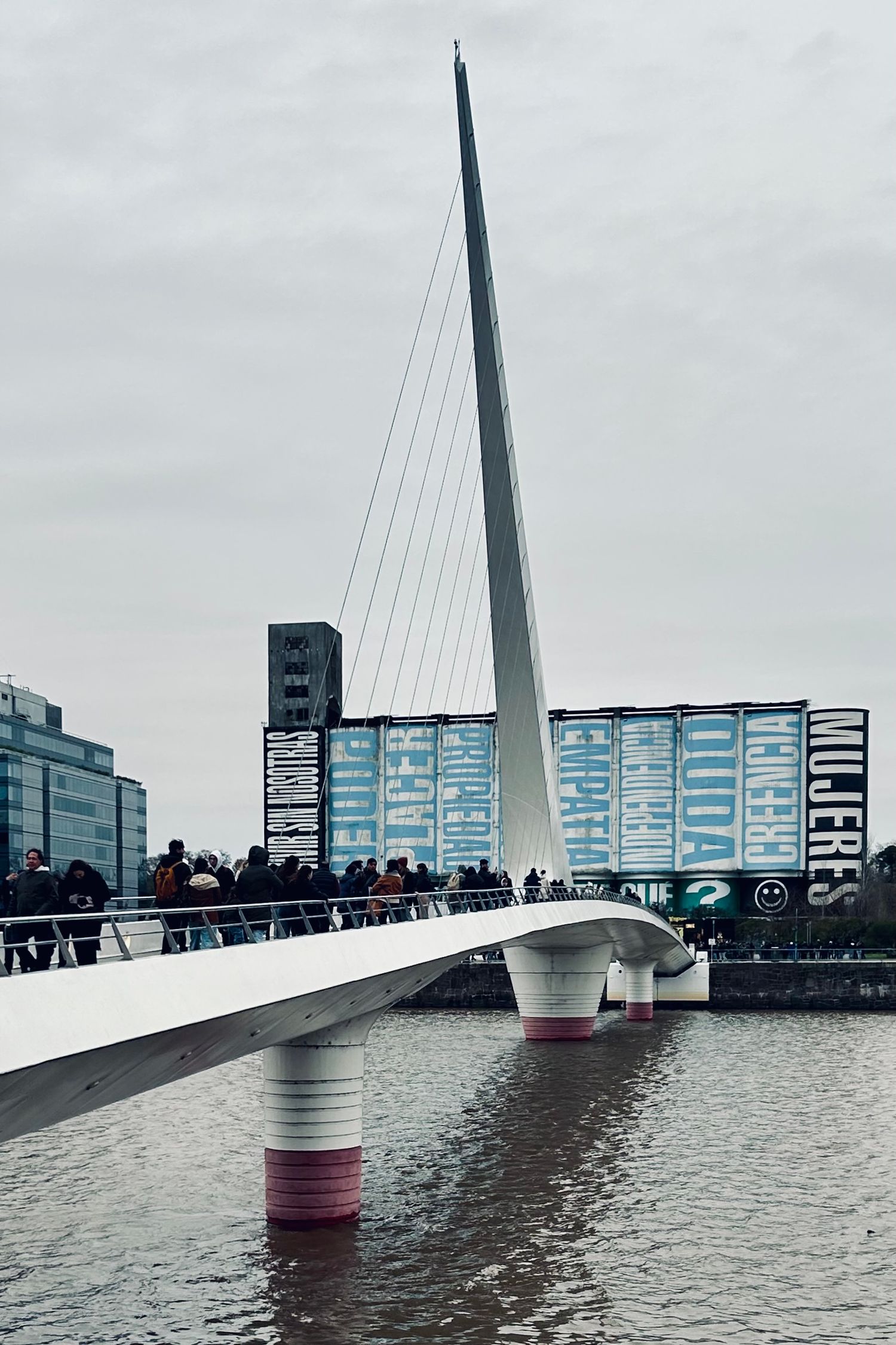 Modern white pedestrian bridge with curved supports spans across calm water with urban buildings in background.