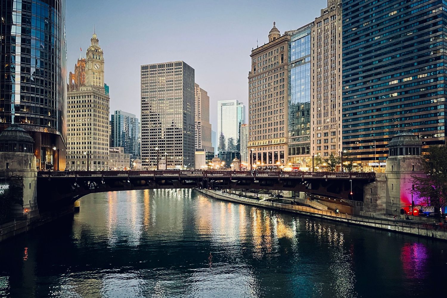 Panoramic cityscape of Chicago River at dusk with illuminated skyscrapers reflecting on water.