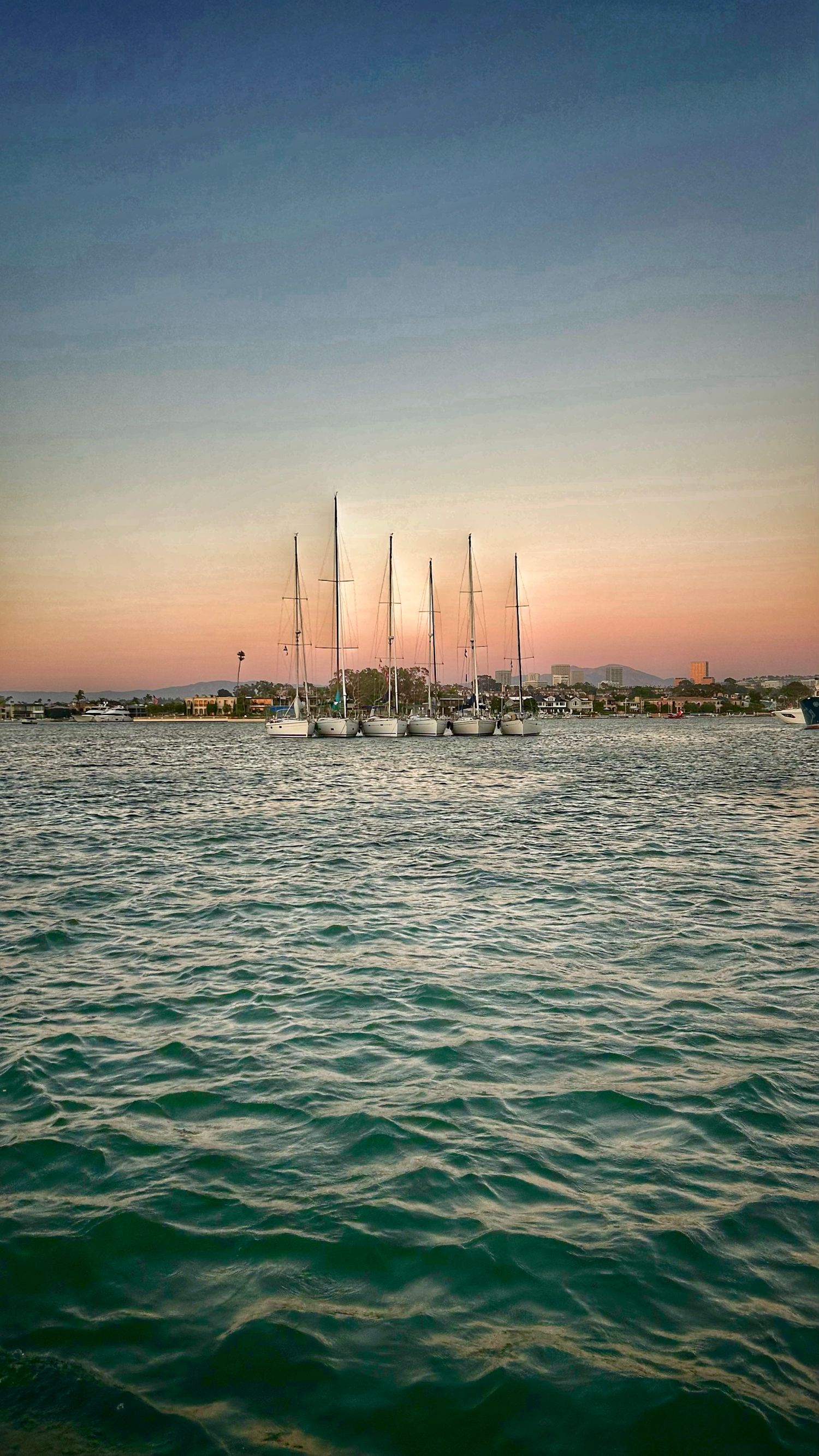 Sailboats anchored in marina at sunset with mountains silhouetted against orange and blue sky.