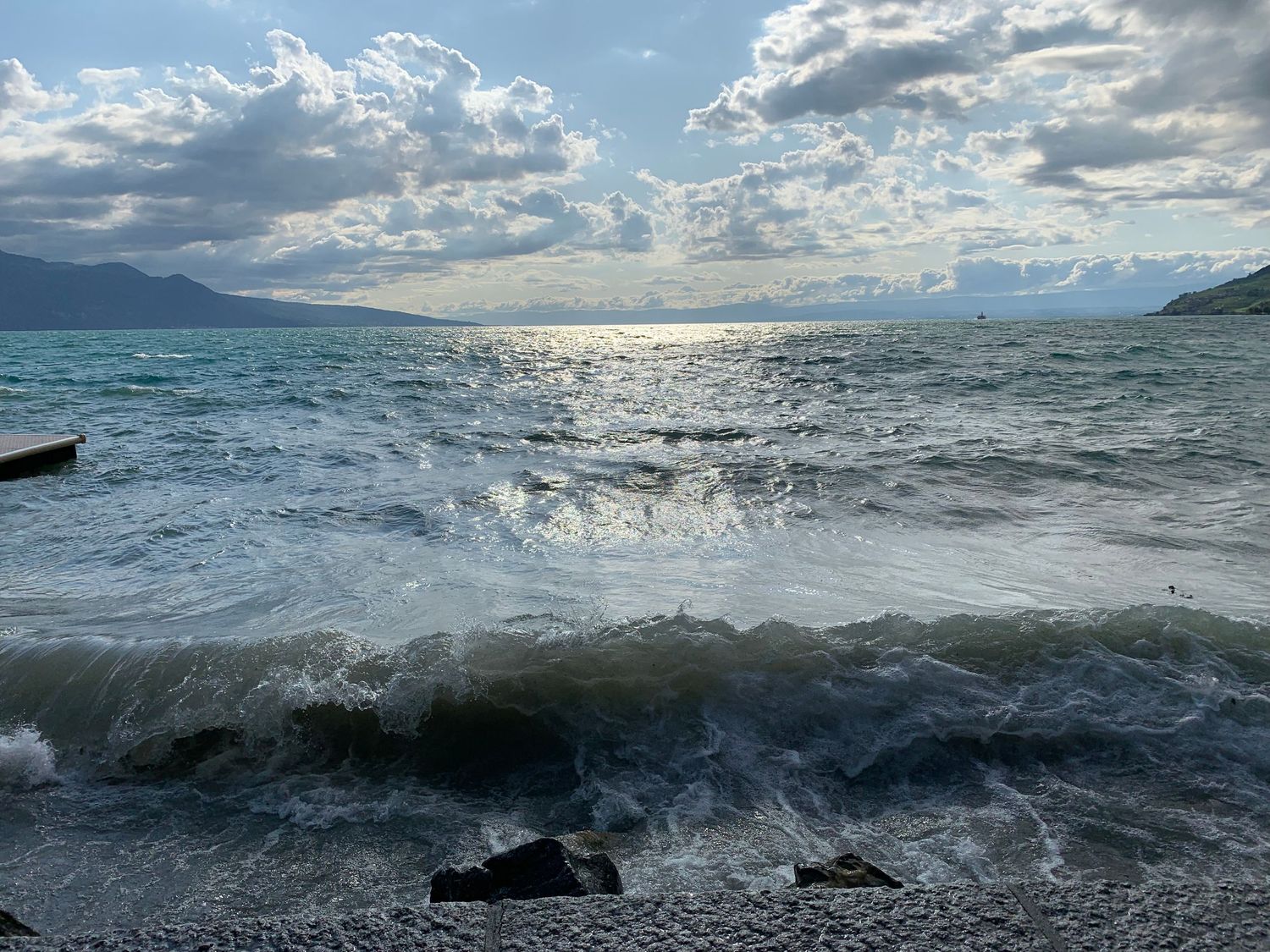Sunlight reflects off turbulent ocean waves with mountains and dramatic clouds on the horizon.