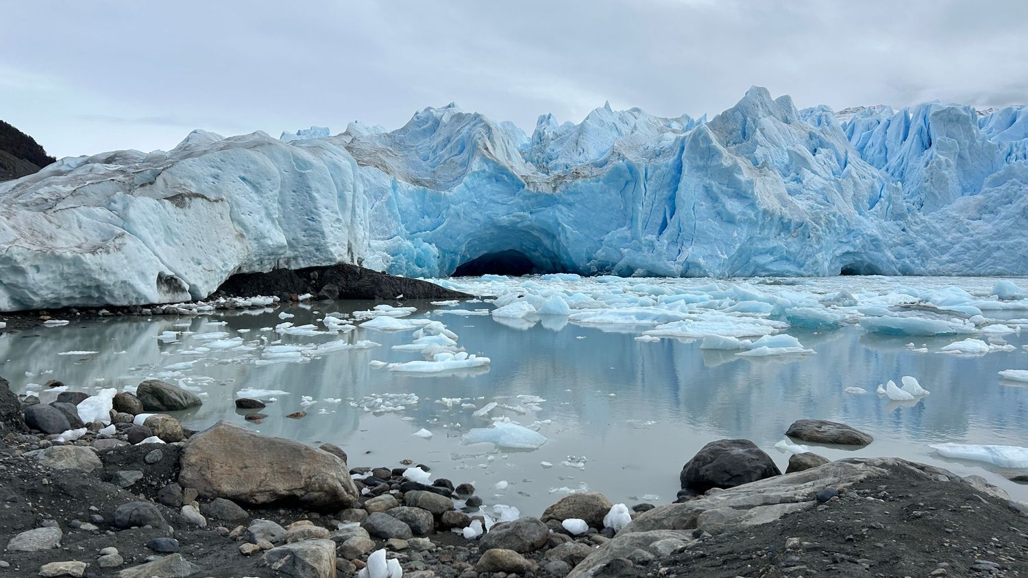Massive blue glacier ice wall reflects in still waters with rocky shore in the foreground.