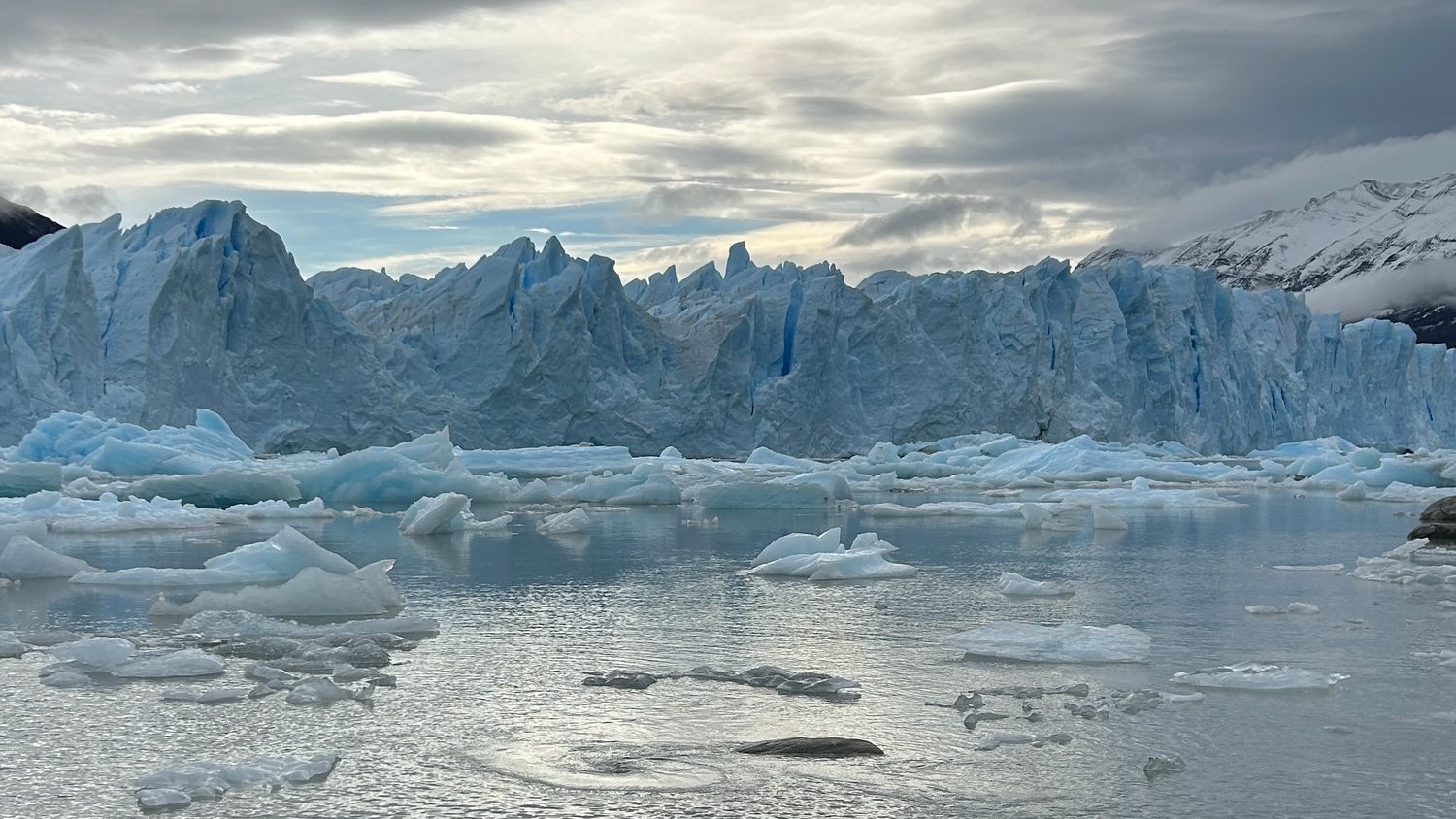 Massive blue glacier ice formations reflect in calm waters under dramatic cloudy skies in a winter landscape.