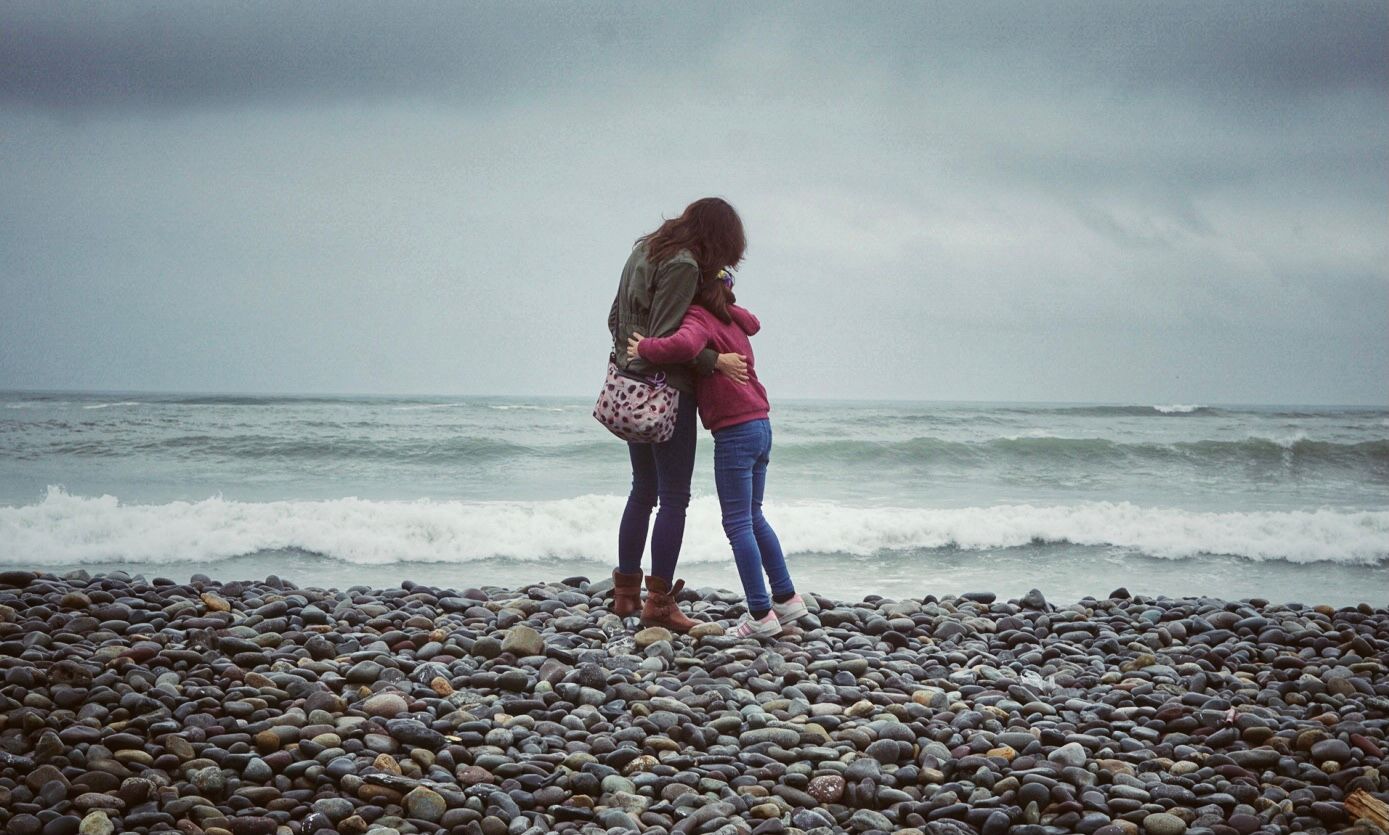 A couple walks hand in hand along a rocky beach with crashing waves and moody overcast skies in the background.