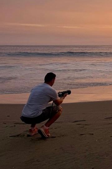 Photographer crouches on sandy beach at sunset capturing ocean waves with camera in golden light.