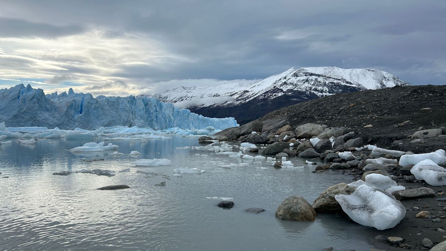 Panoramic view of snow-capped mountains and glaciers reflecting in calm arctic waters under cloudy skies.