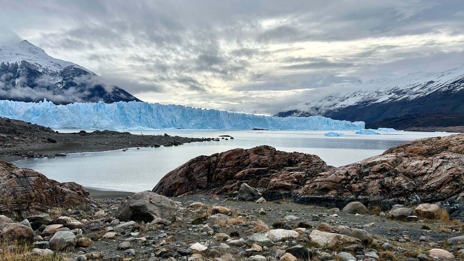 Snow-covered mountains frame a dramatic glacier landscape with rocky outcrops under cloudy skies.