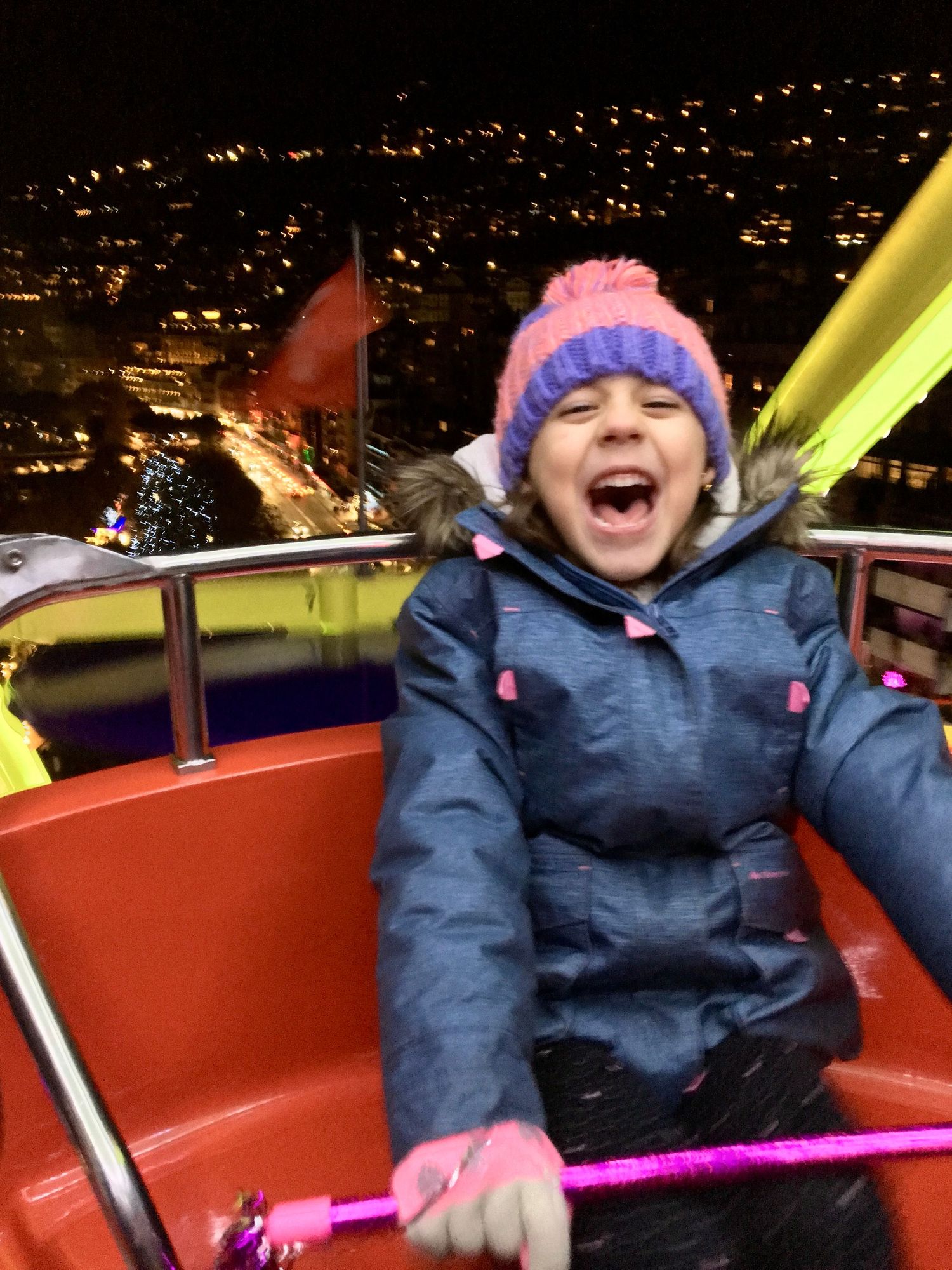 A child wearing a blue winter coat and pink hat expresses pure joy while riding on an amusement park ride at night.