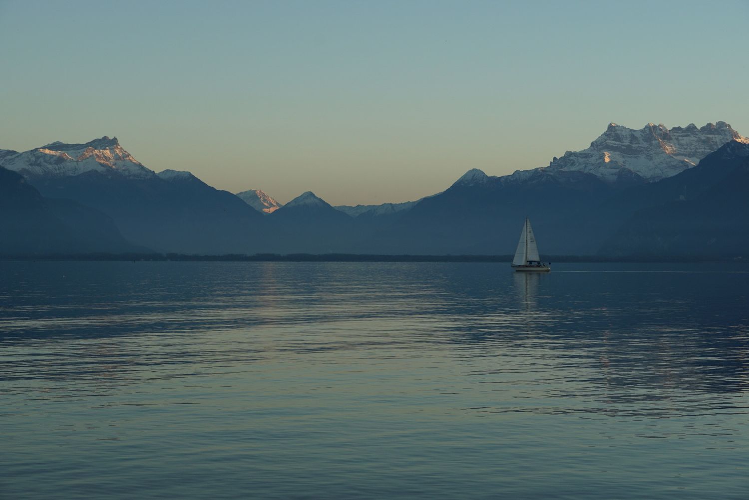 Sailboat glides across tranquil blue waters with mountain silhouettes against a serene sunset sky.
