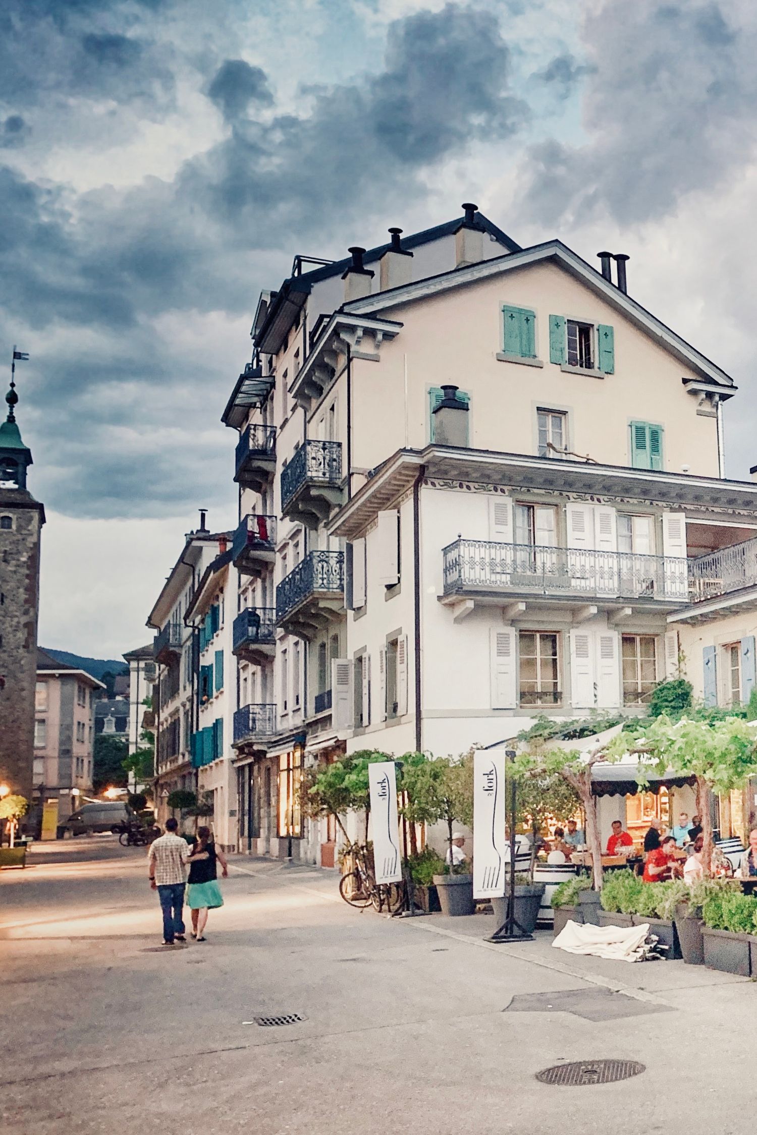 Quaint European-style buildings and shops line a cobblestone street under dramatic cloudy skies.