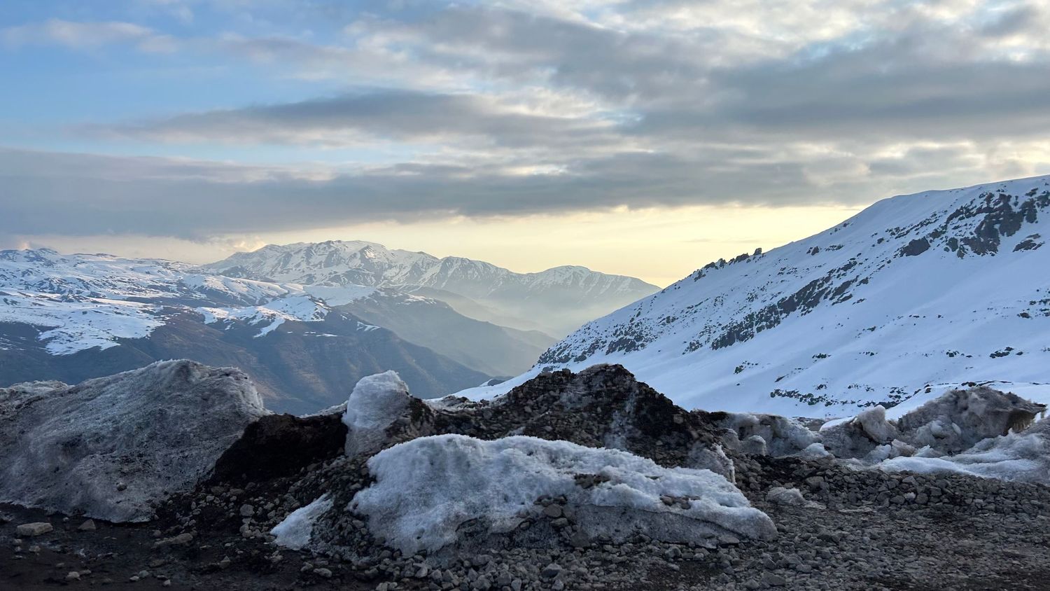 Snow-covered mountain peaks and valleys under dramatic cloudy skies during sunset in a winter landscape.