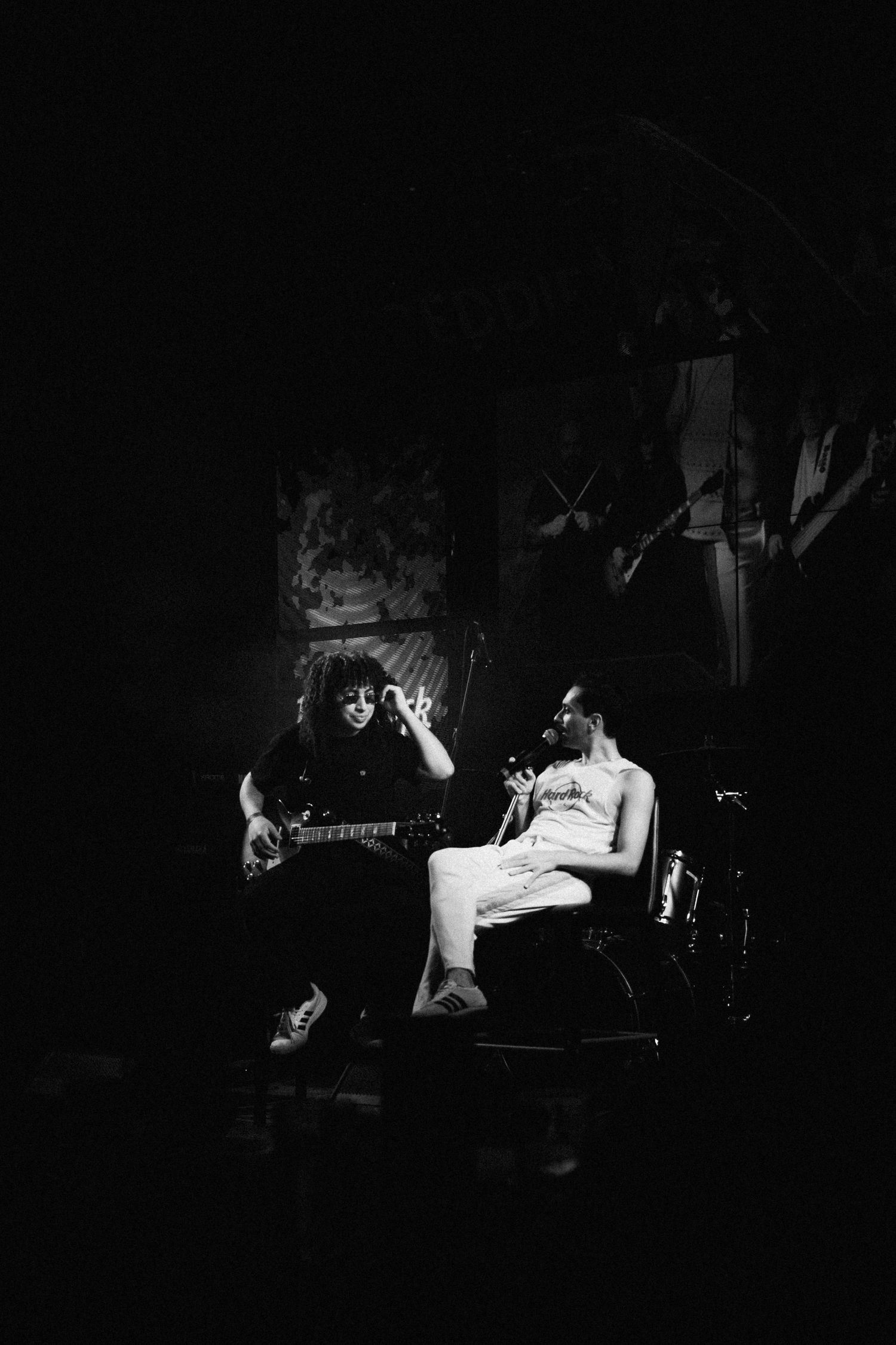 Plano aberto do palco escuro, com guitarrista em pé à esquerda e o vocalista sentado à direita, em preto e branco.