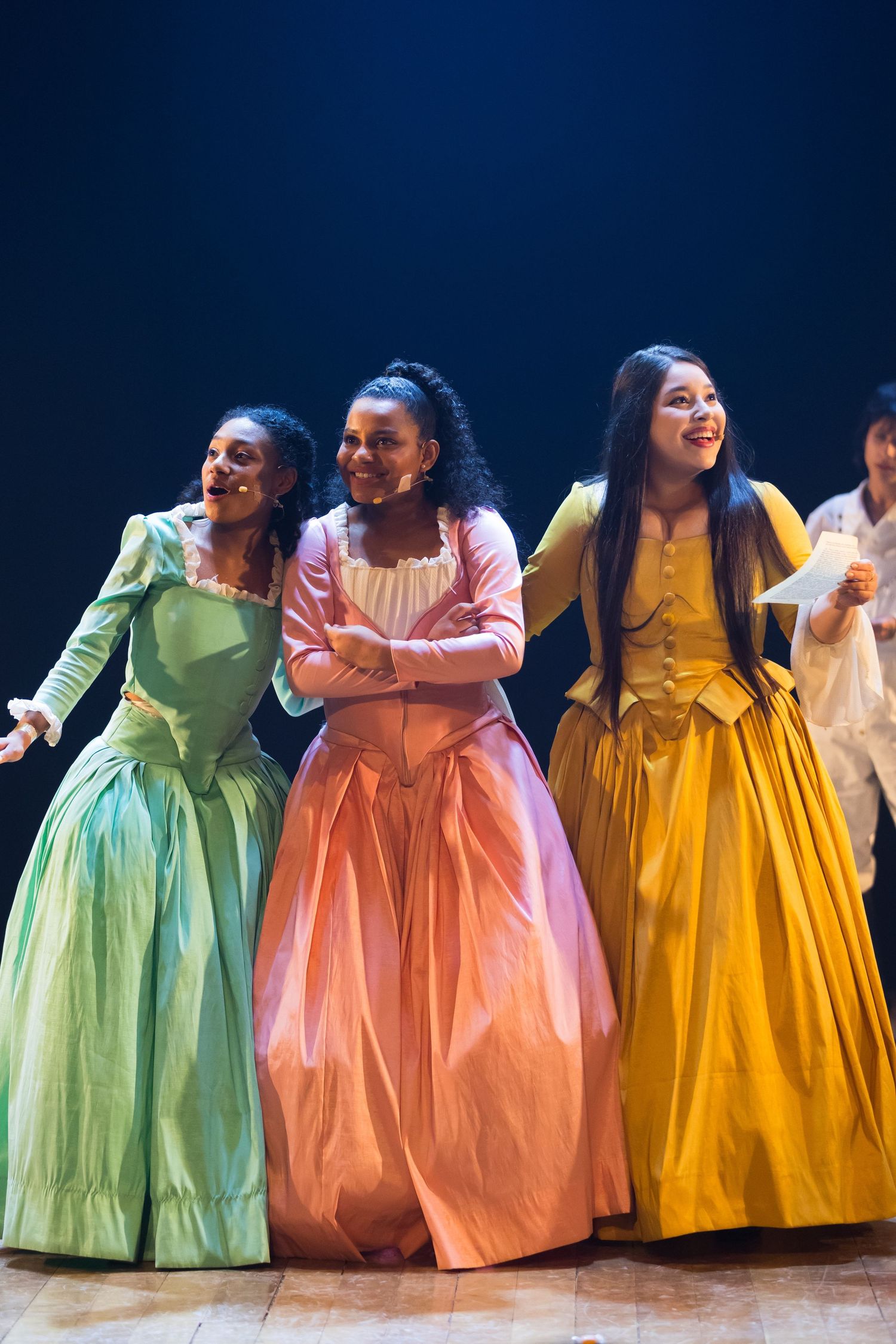Três intérpretes em vestidos de época verde, rosa e amarelo lado a lado, com o elenco ao fundo.